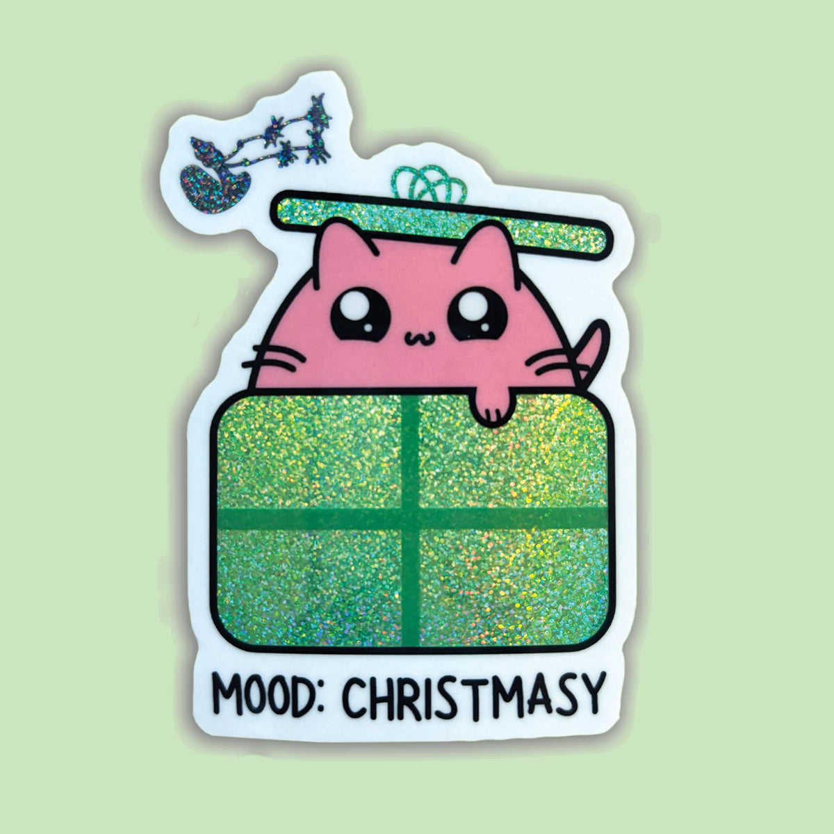 A die-cut sticker featuring a cute pink cartoon cat peeking over a glittery green Christmas gift box, with a small sleigh and stars above and the text &