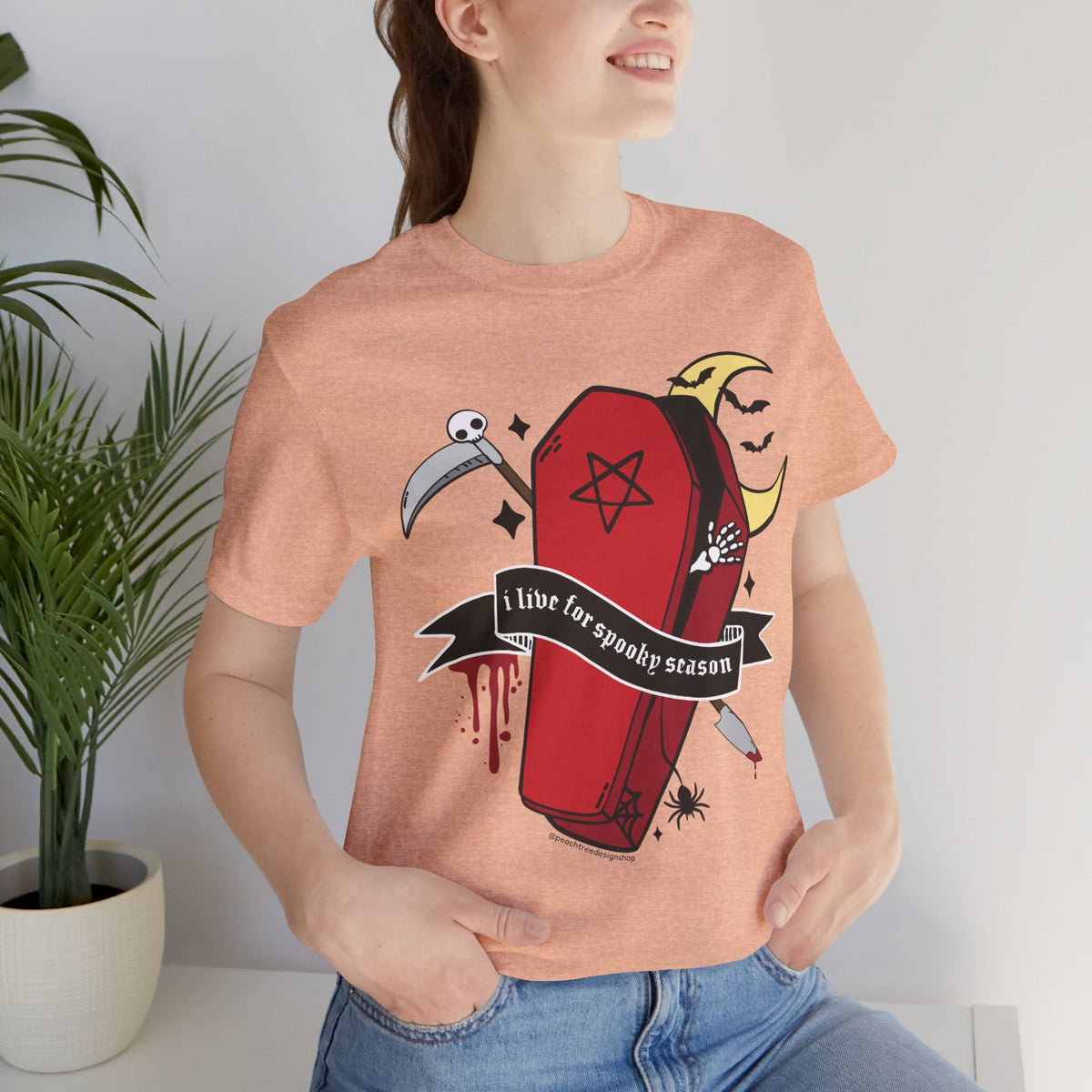 A woman wearing a heathered peach t-shirt with a gothic graphic of a red coffin, a scythe, a spider, and a banner that reads &
