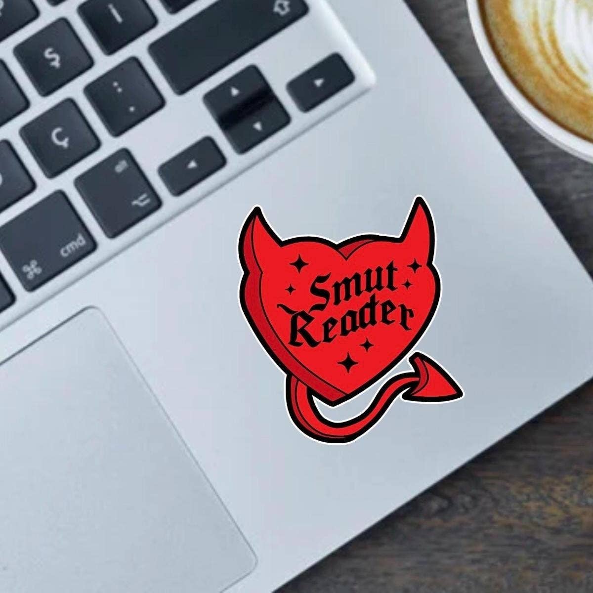 Red heart-shaped devil sticker with horns and tail reading “Smut Reader” on a laptop next to a cup of coffee.