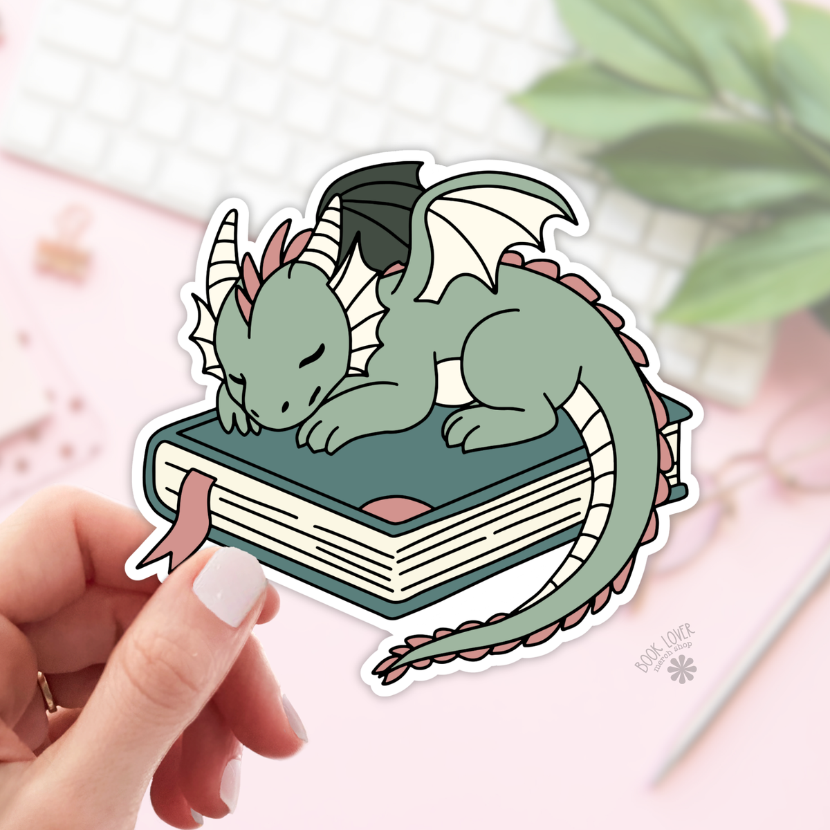 Sticker of a green dragon on a book held by a hand with a blurred background