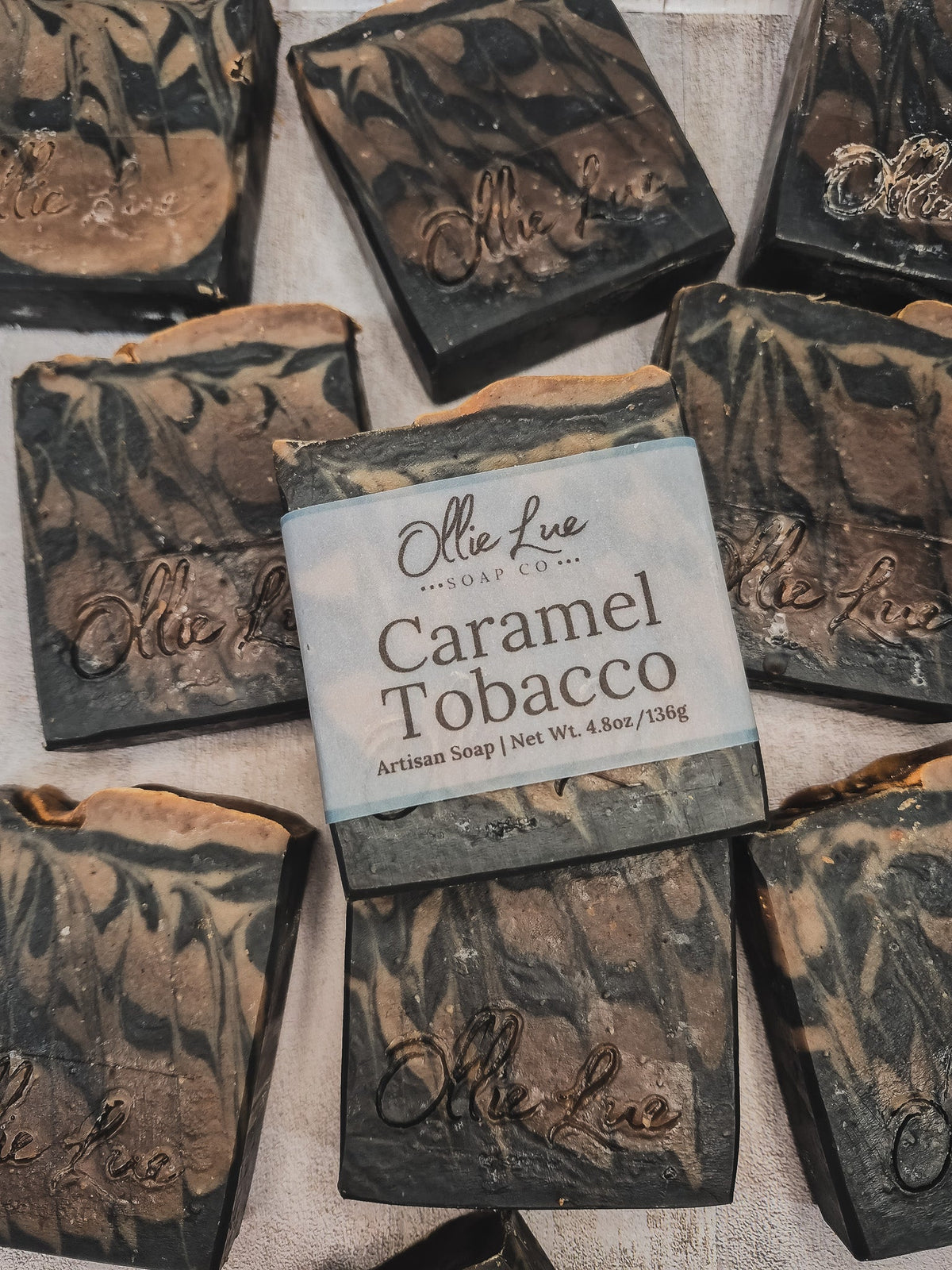 Several handmade soap bars are arranged on a light fabric background. The center soap is labeled &