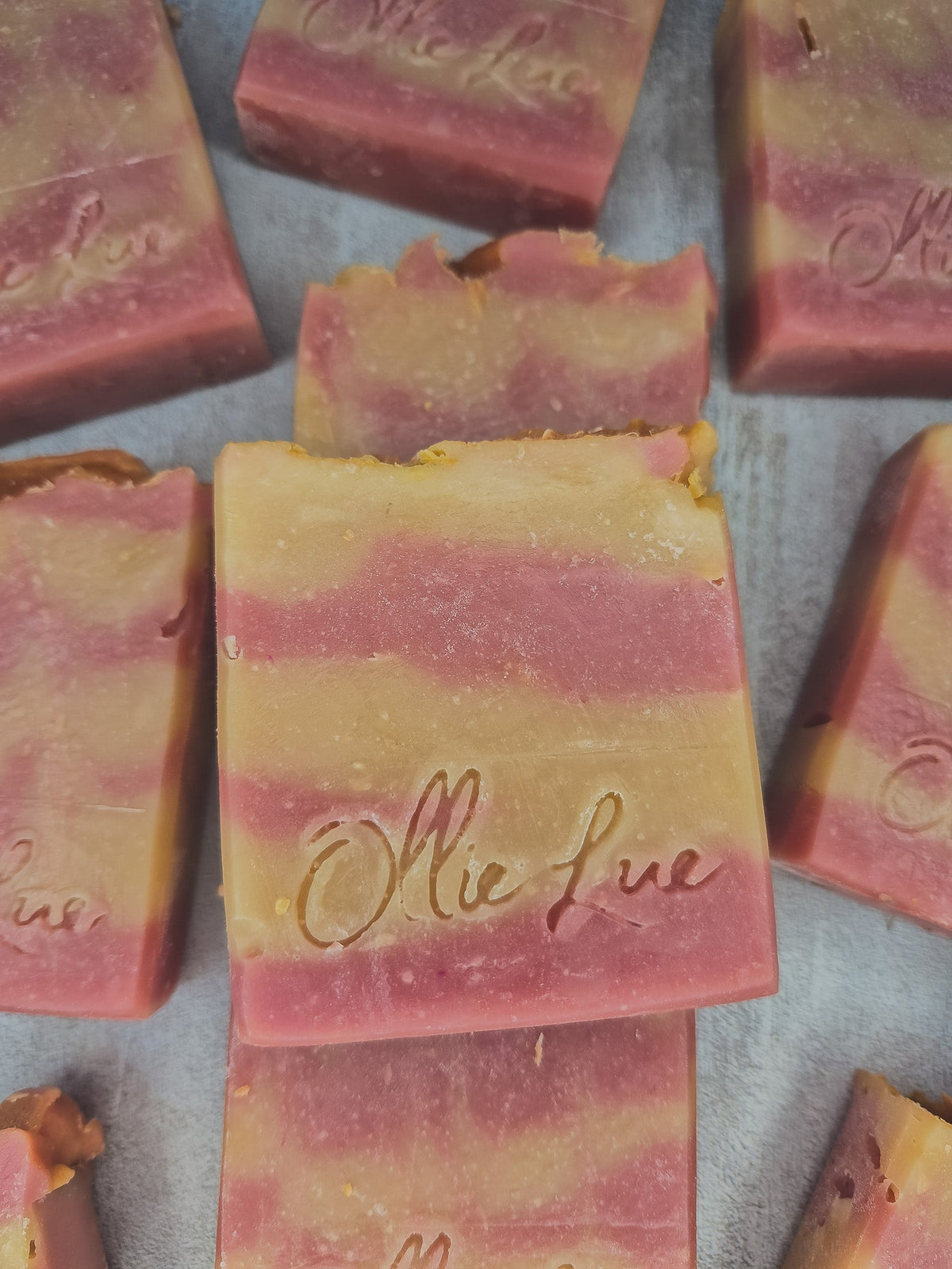 Several handmade rectangular bars of soap in pink and pale yellow marbled layers, arranged on a light gray surface. The center bar is upright and embossed with the brand name &
