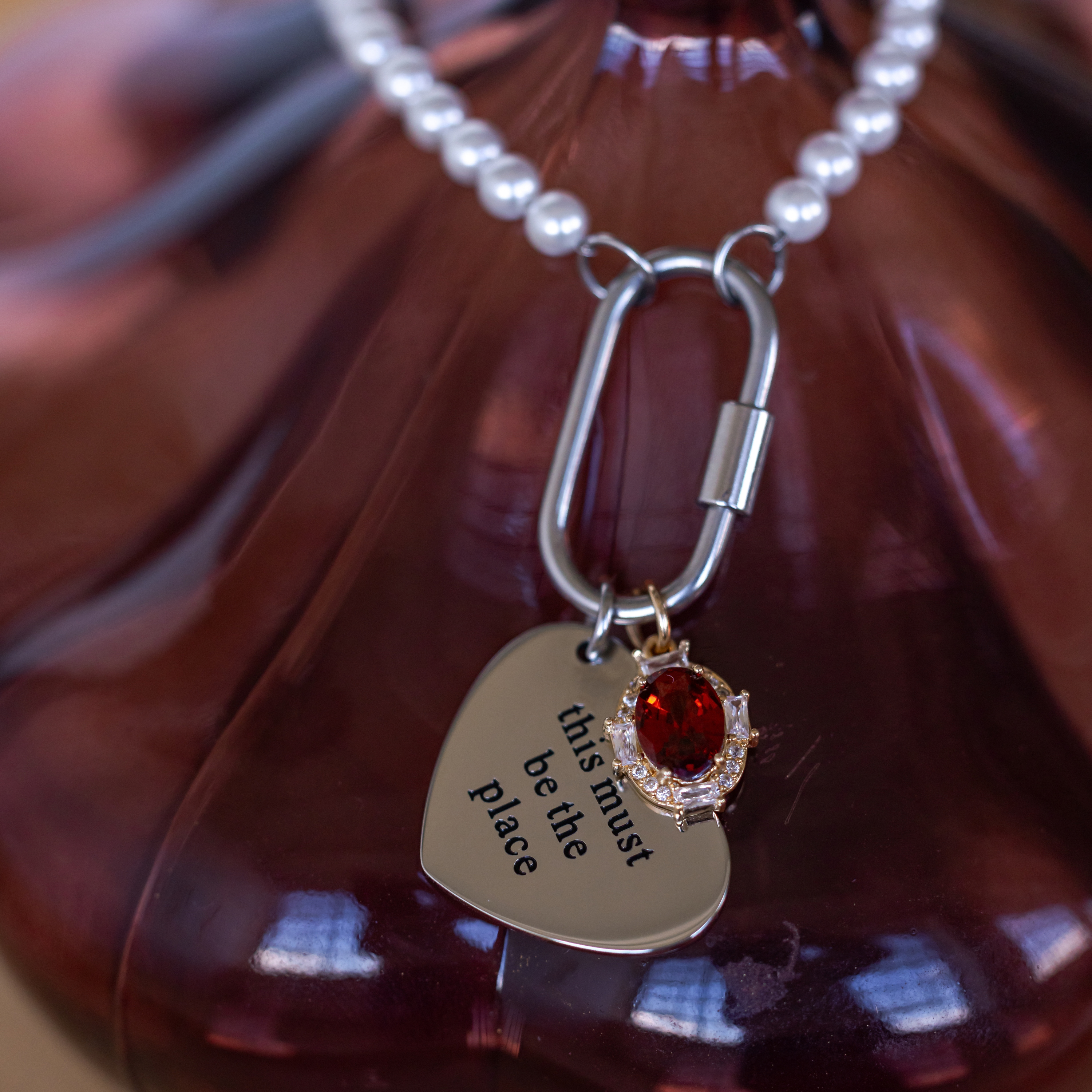 Heart-shaped pendant with engraved text and red gemstone on a chain against a brown background