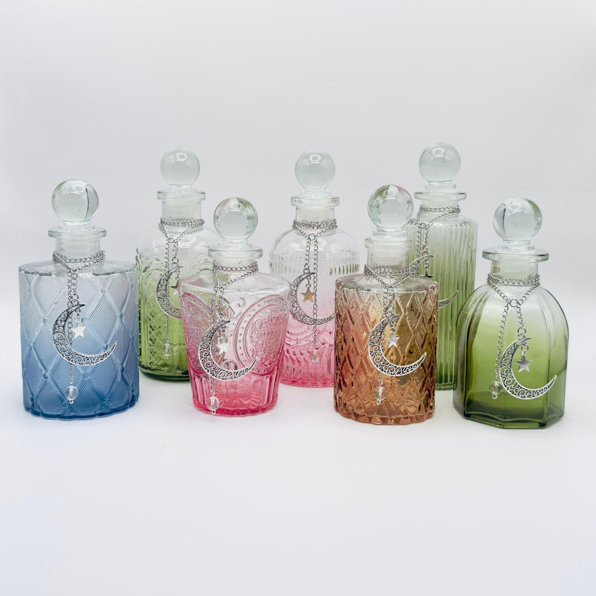 A set of eight decorative glass decant bottles with stoppers, featuring a silver crescent moon and star motif. The bottles are shown in various colors including blue, green, pink, orange, and yellow, arranged on a light gray background.