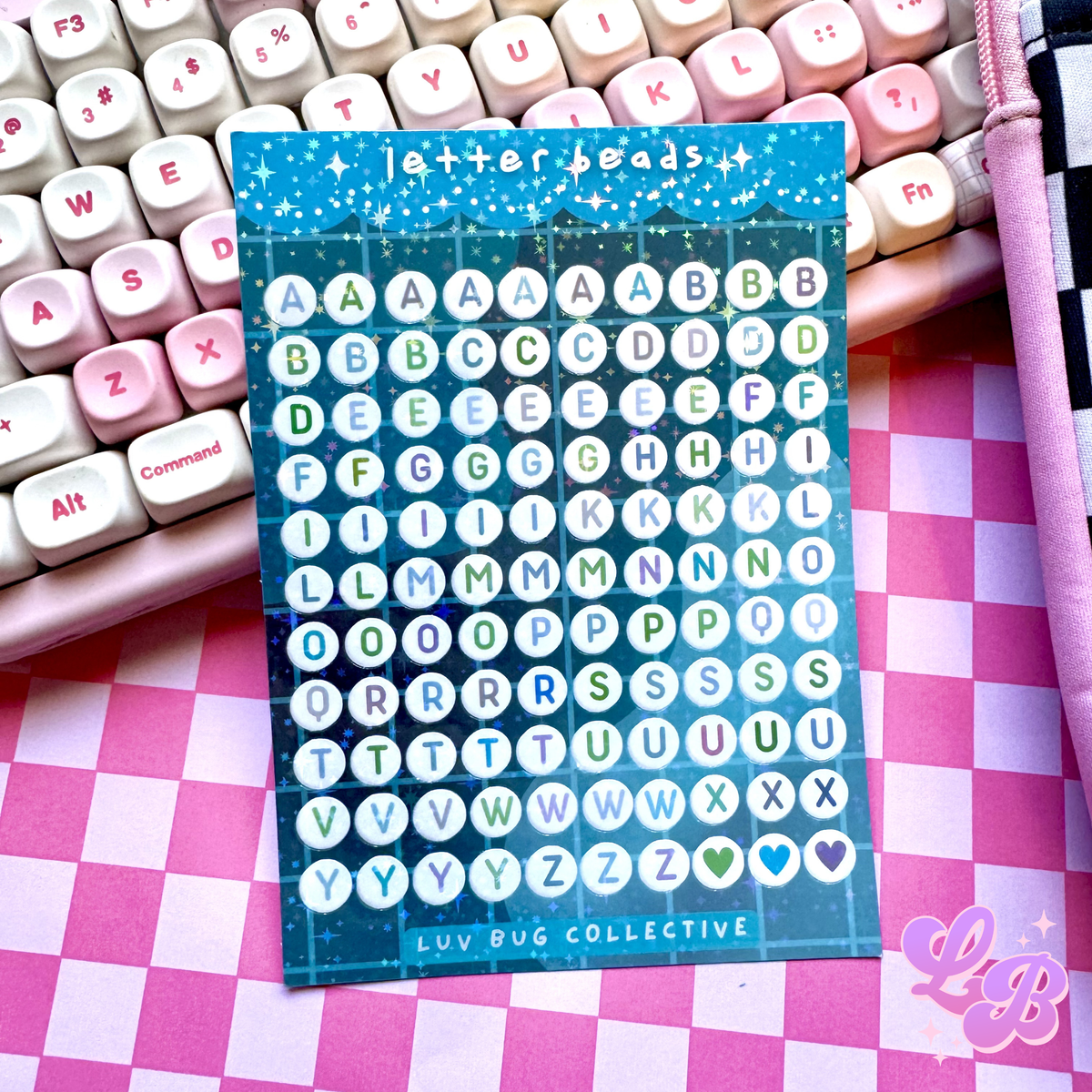 Sticker sheet of blue, purple, and green toned letter beads resting on a pink keyboard and pink checkered mat in the background