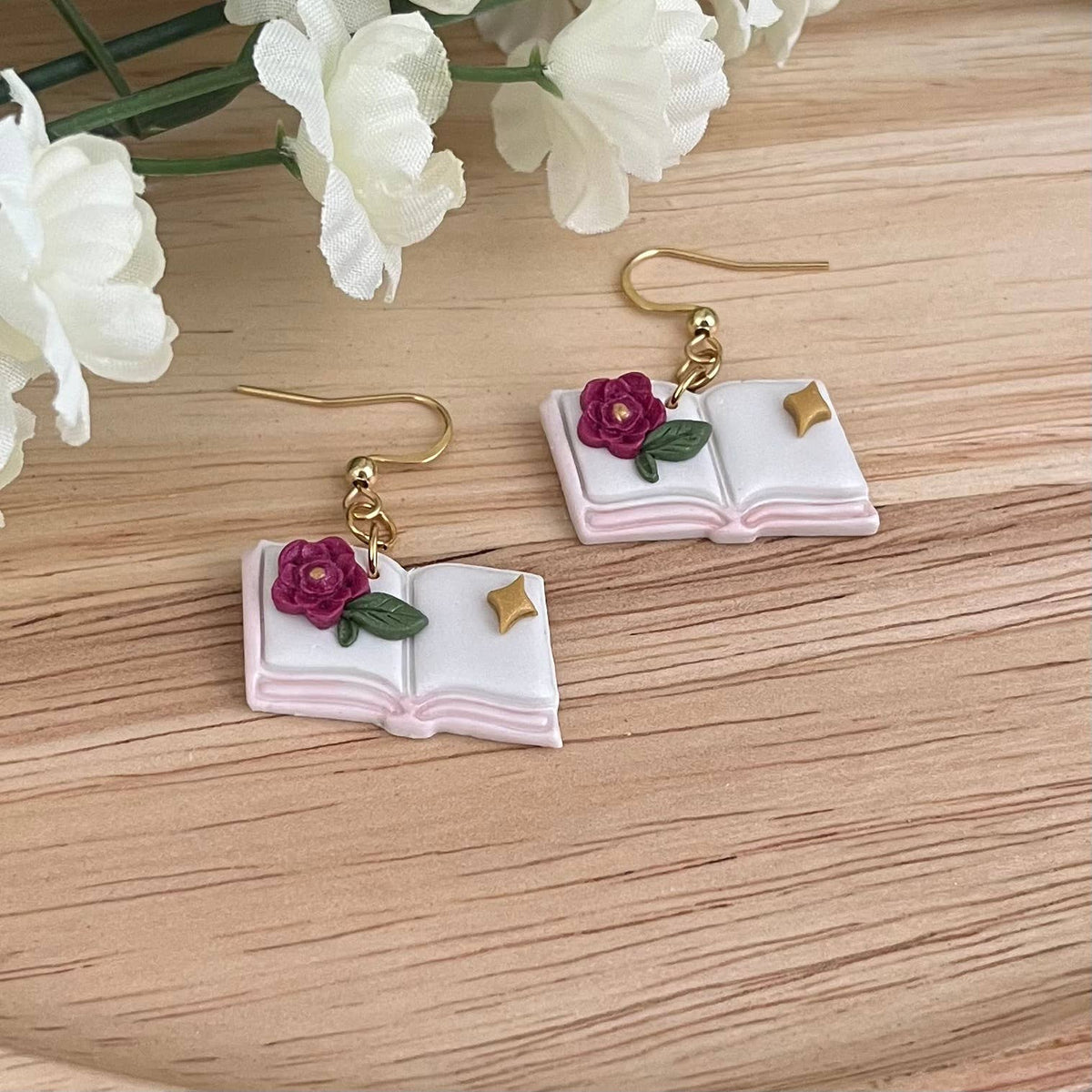 Book-shaped earrings with floral details on a wooden surface with white flowers.
