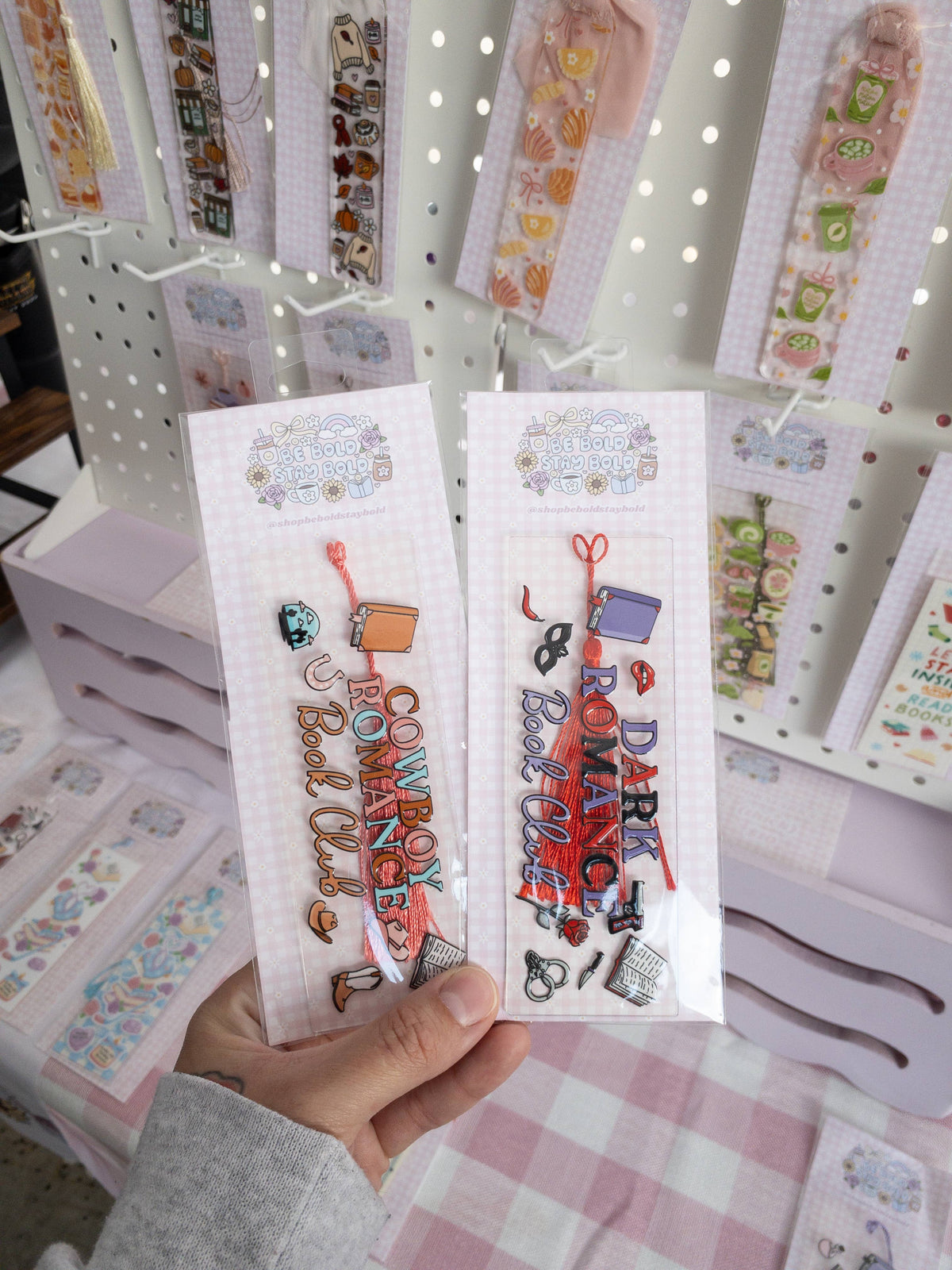 Two bookmarks with colorful text and designs held by a hand, with a display of more bookmarks in the background.