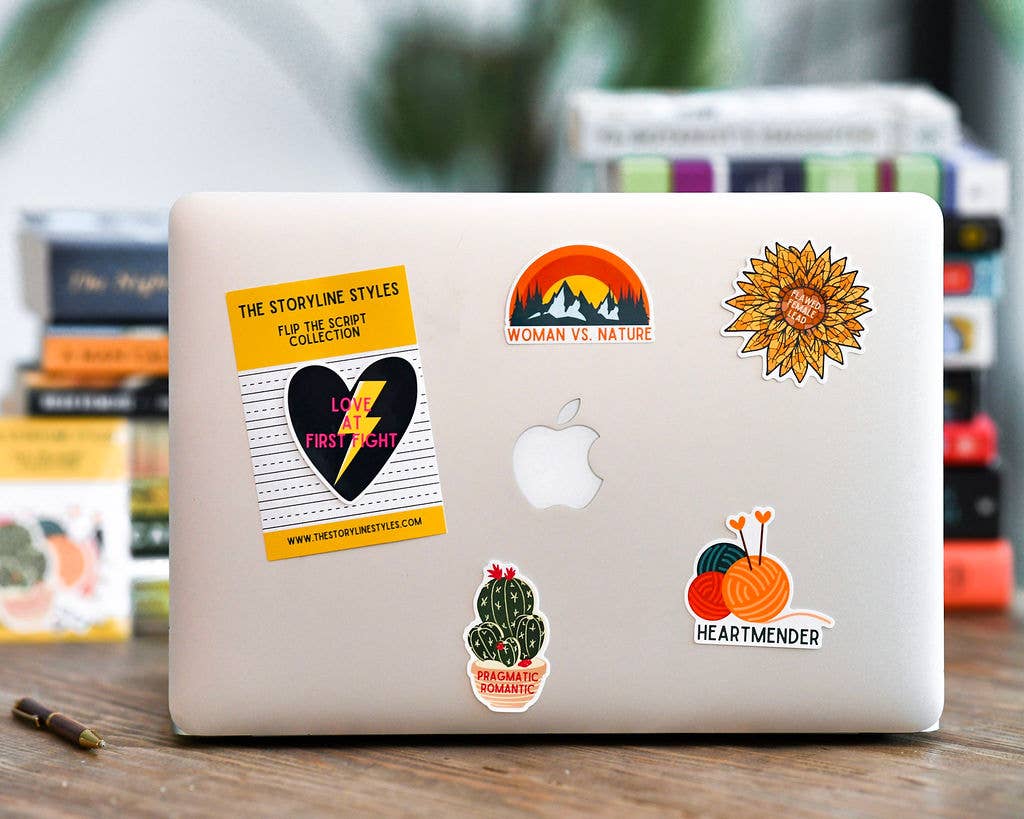 Laptop with various stickers on a wooden surface with books in the background