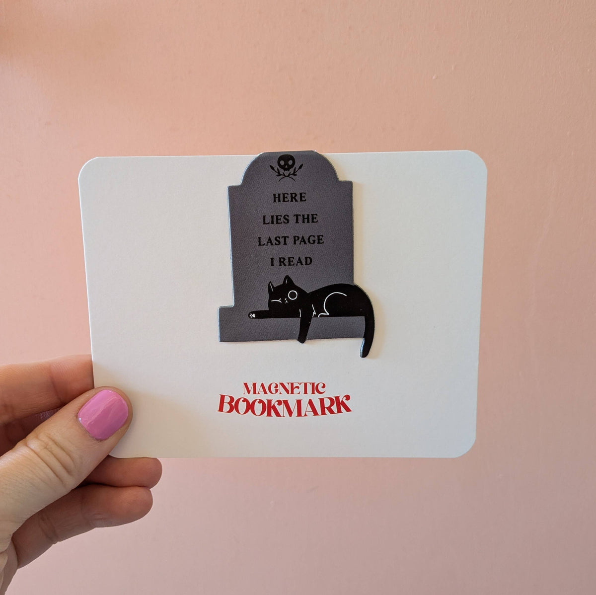 Magnetic bookmark with cat design and text held by a hand on a pink background