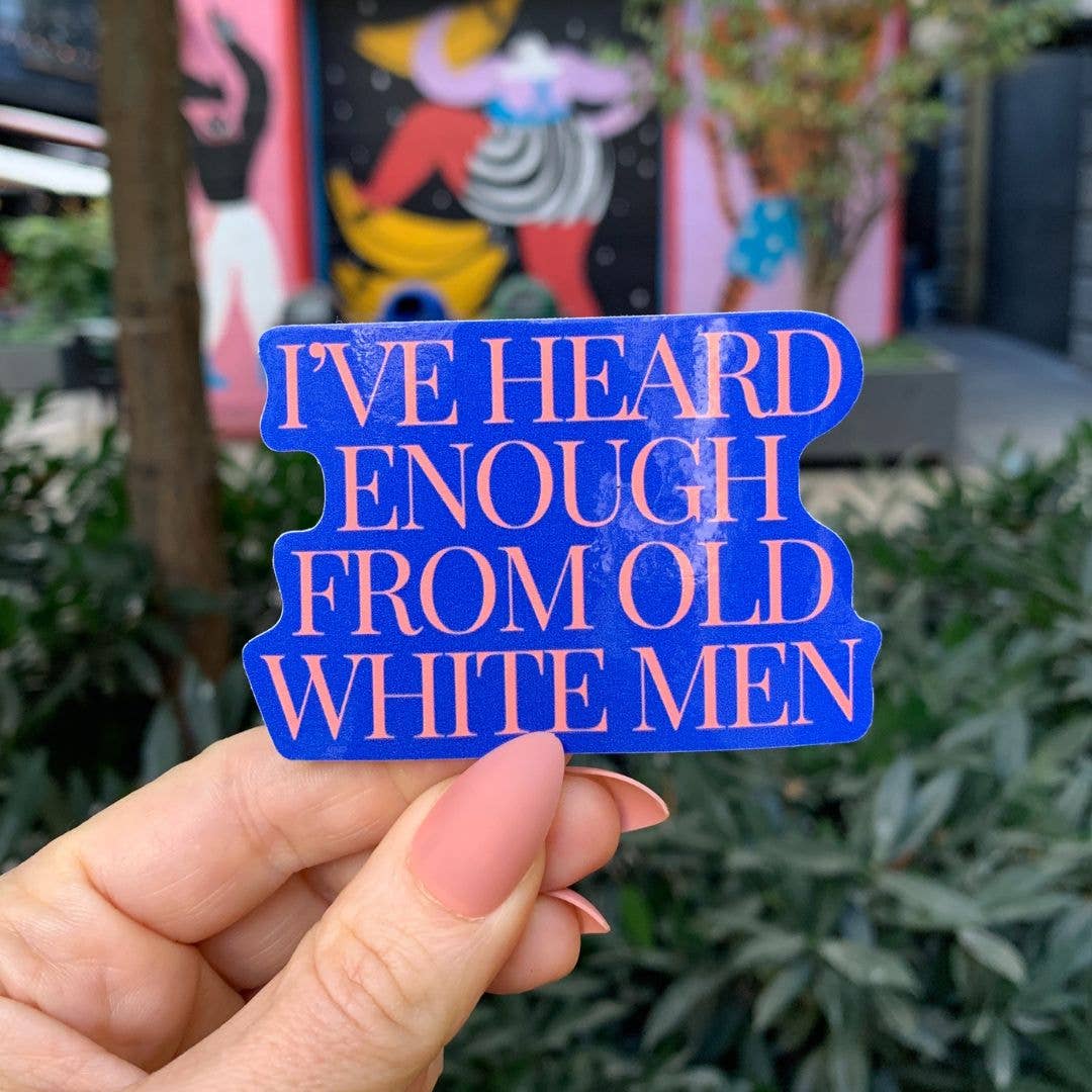 Blue sticker with text held by a hand in front of a colorful mural.