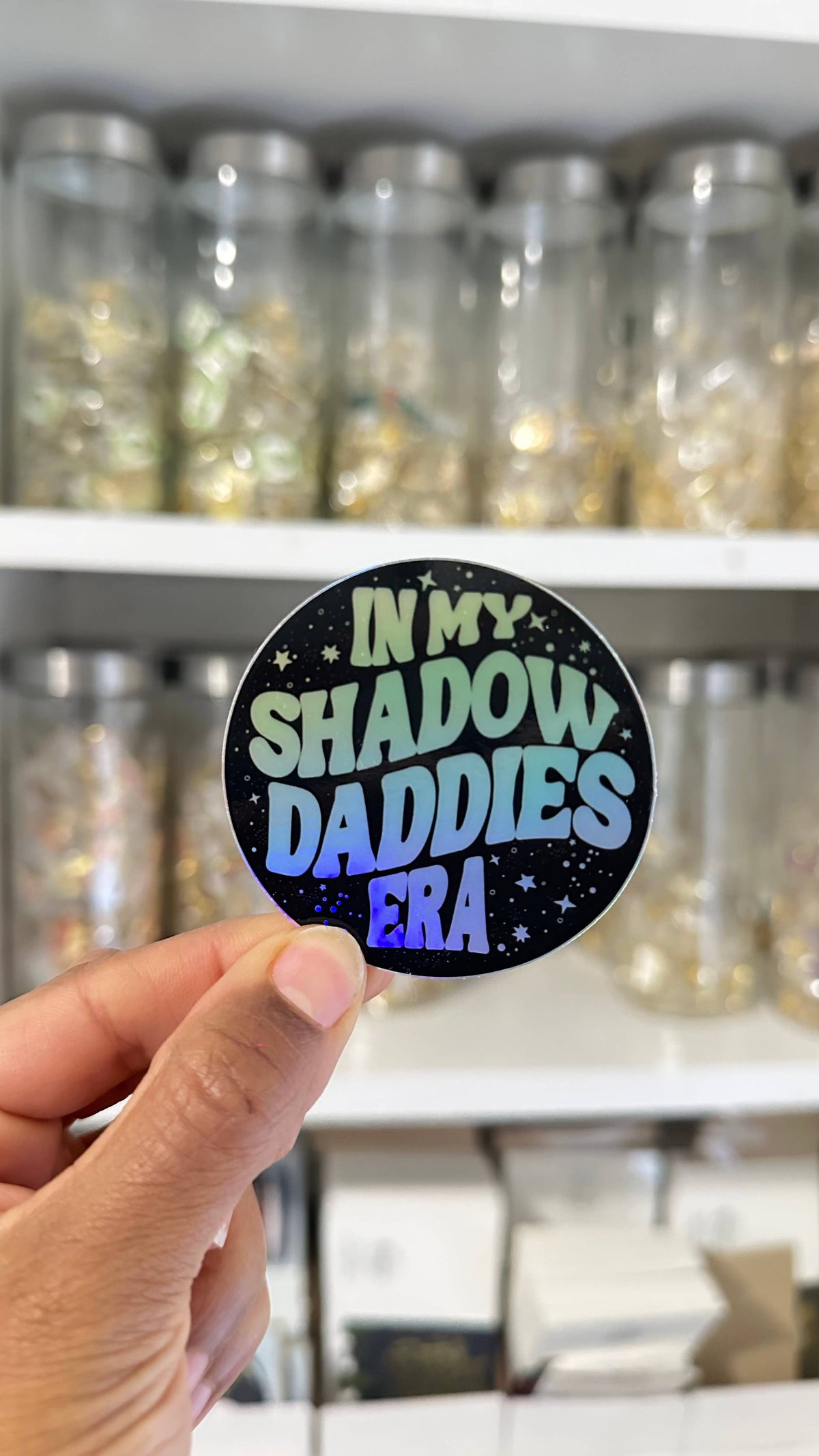 Person holding a round sticker with text in front of shelves with jars.
