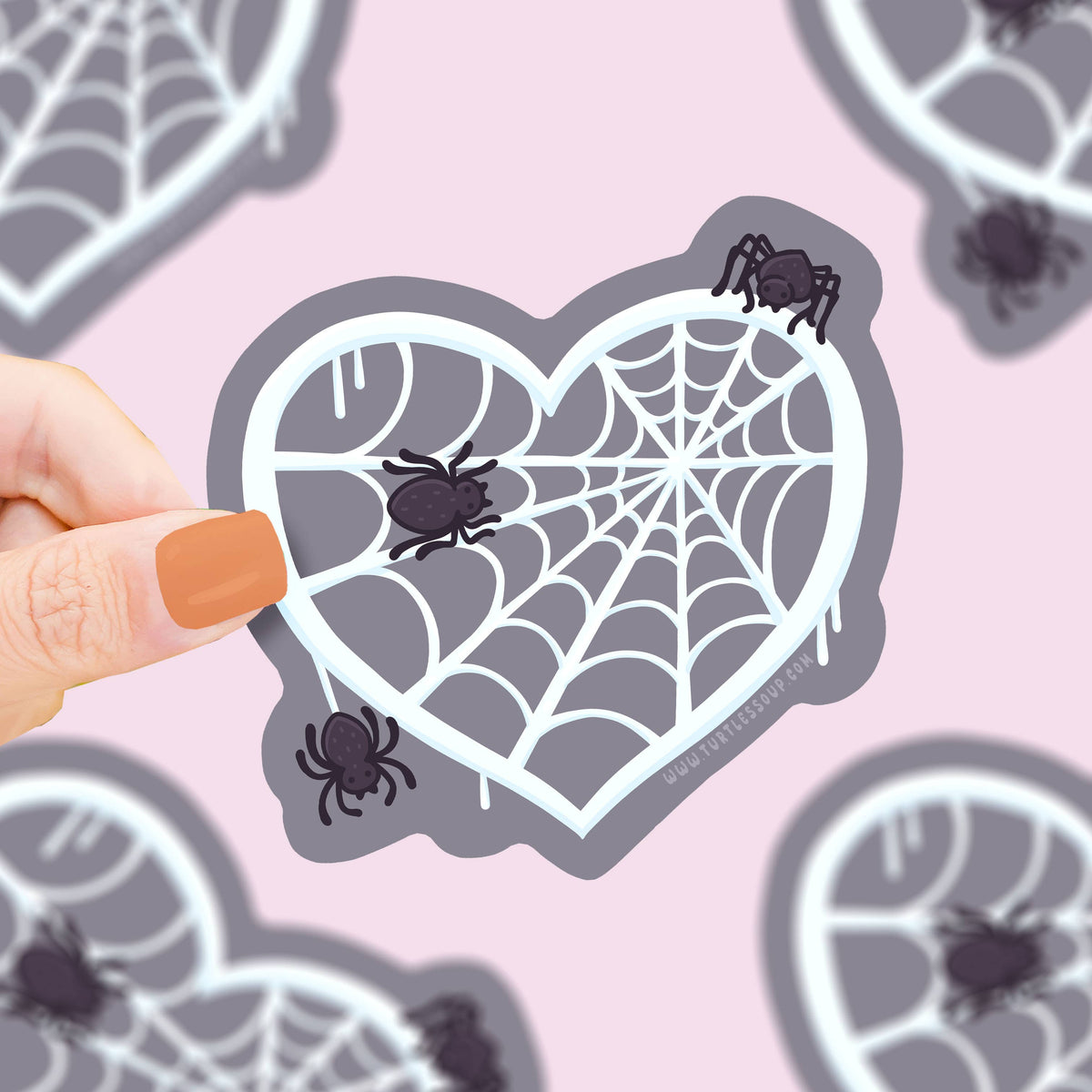 Heart-shaped spider web sticker held by a hand on a pink background with more stickers.