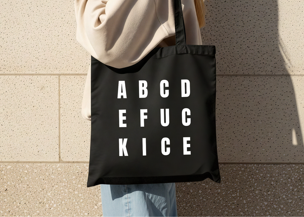 Person holding a black tote bag with white text reading ABCDE FUCK ICE against a beige wall.