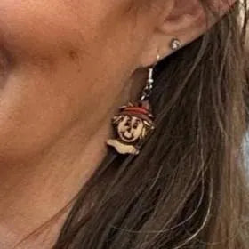 Close-up of a person wearing hand-painted wooden scarecrow dangle earrings with smiling scarecrow faces, straw hair, and burgundy hats on hypoallergenic .925 sterling silver hooks, highlighting the lightweight size and detailed design.
