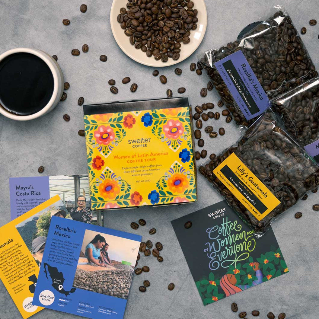 Overhead shot of a Coffee Tour box featuring women of Latin America; three coffee bags, with three info cards, a cup of coffee and beans scattered on a gray surface