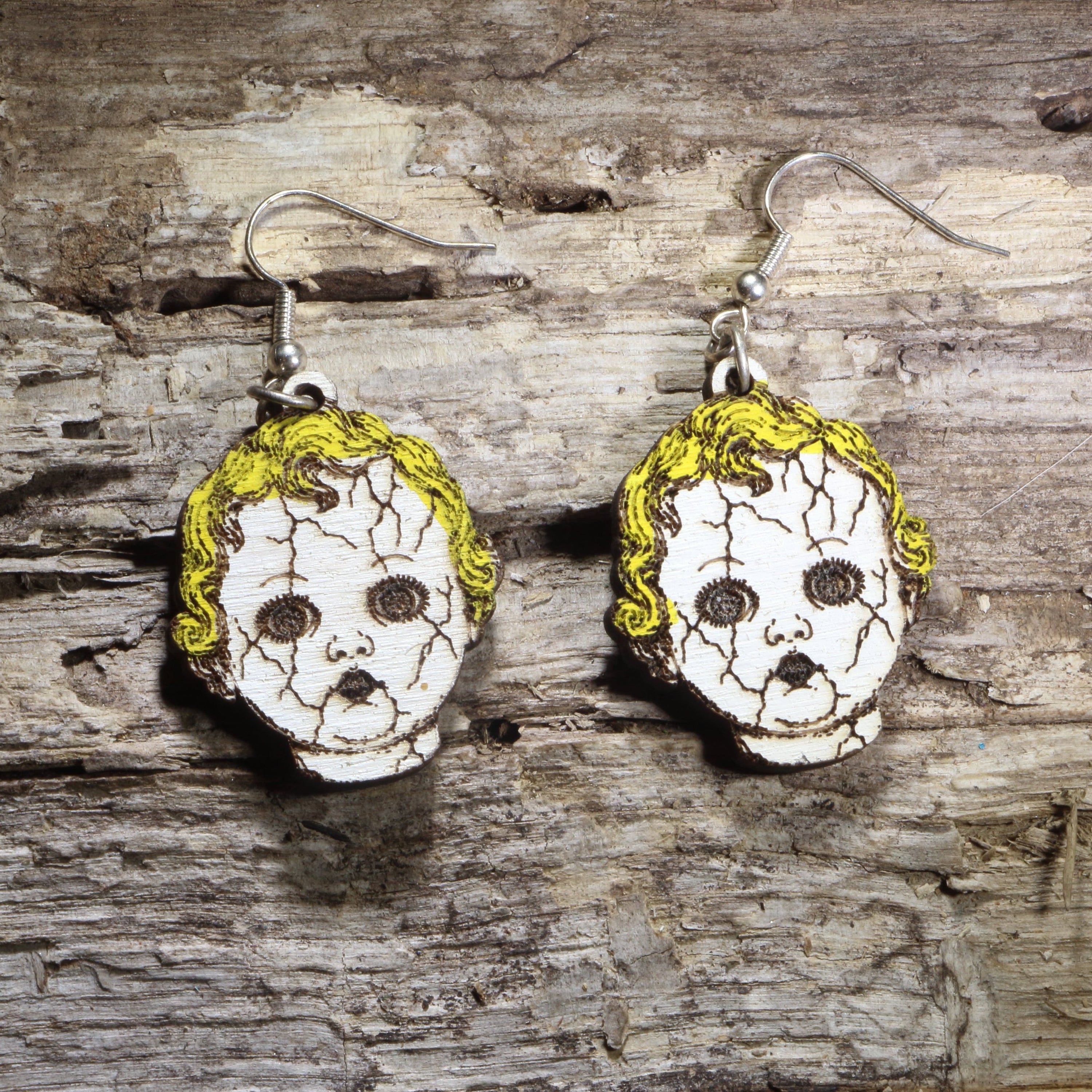 Handcrafted creepy cracked doll head wooden dangle earrings featuring vintage porcelain-style doll faces with cracked details and eerie expressions. Lightweight laser-cut wooden earrings with hypoallergenic .925 sterling silver hooks, perfect for Halloween lovers, horror fans, haunted house actors, gothic fashion, creepy doll collectors, and spooky statement jewelry.
