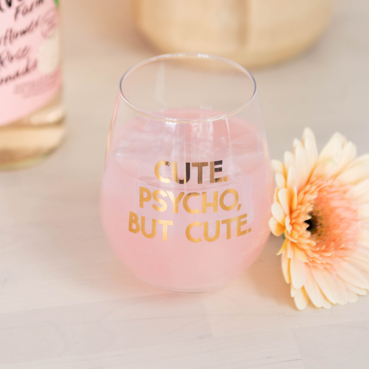 "Cute. Psycho, But Cute." stemless glass with a pink drink beside a bottle and a peach daisy.