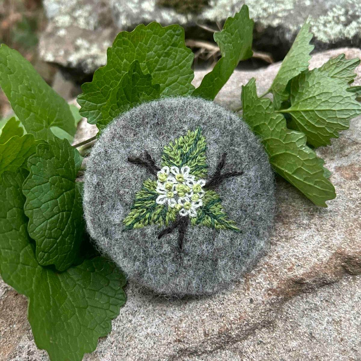Embroidered Garlic Mustard motif on grey wool felt, surrounded by garlic mustard leaves on stone