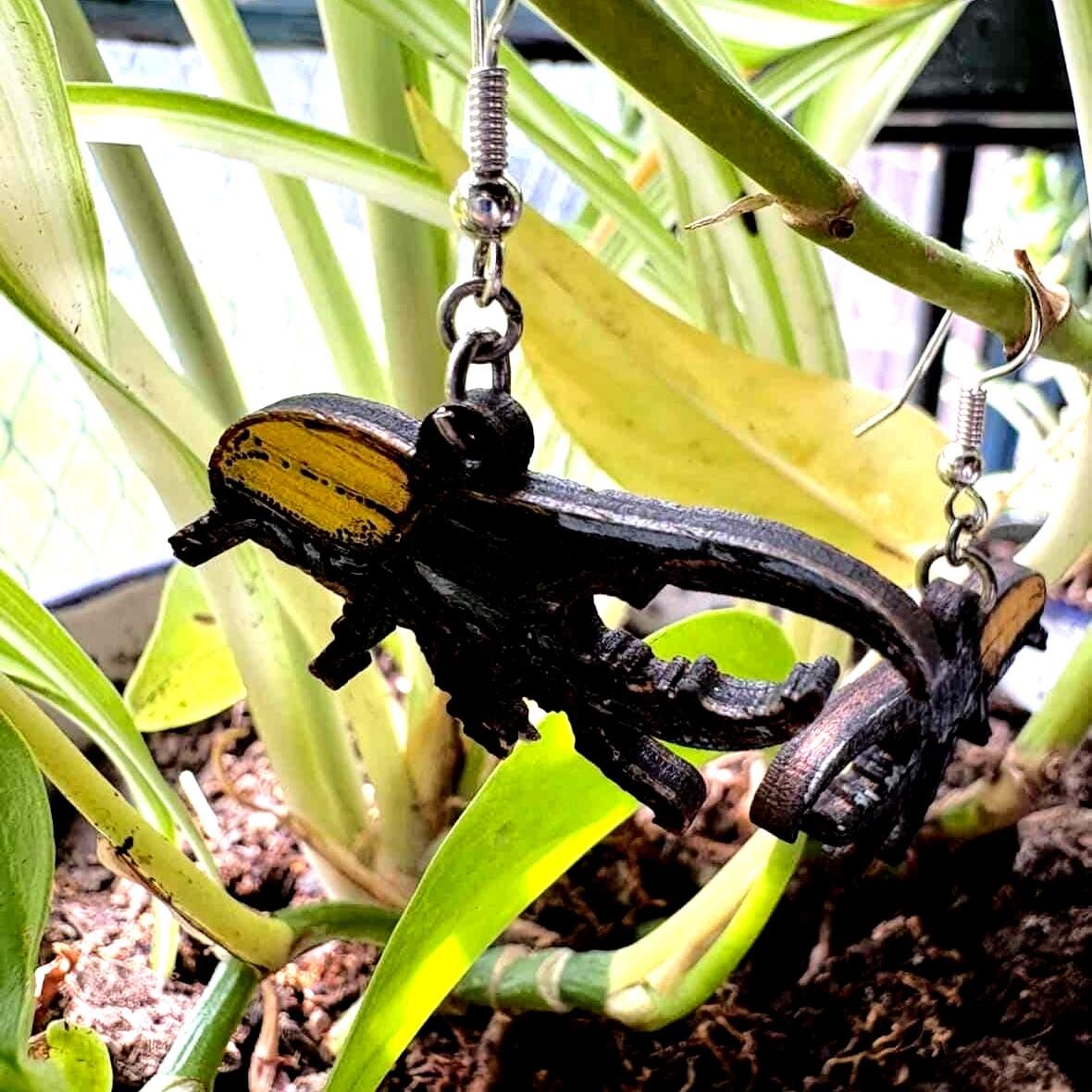 Hand-painted wooden stag beetle dangle earring with black body and golden wing covers hanging from a green houseplant. Lightweight insect jewelry with hypoallergenic .925 sterling silver hook.