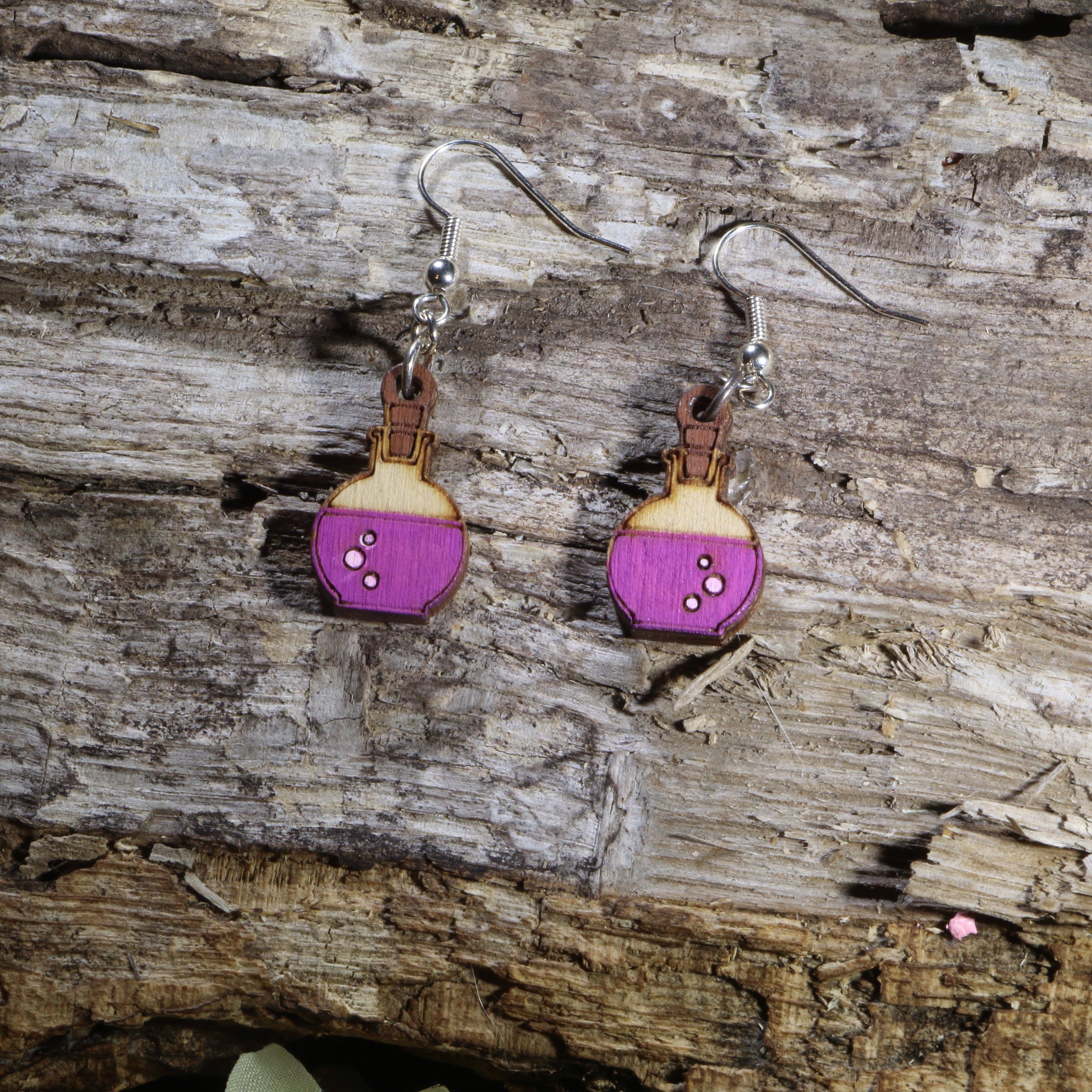 Handcrafted wooden potion bottle dangle earrings featuring vibrant purple potion details and engraved accents. These lightweight fantasy-inspired earrings are finished with hypoallergenic .925 sterling silver hooks, making them the perfect accessory for gamers, fantasy fans, witches, alchemists, tabletop RPG players, and lovers of magical jewelry.