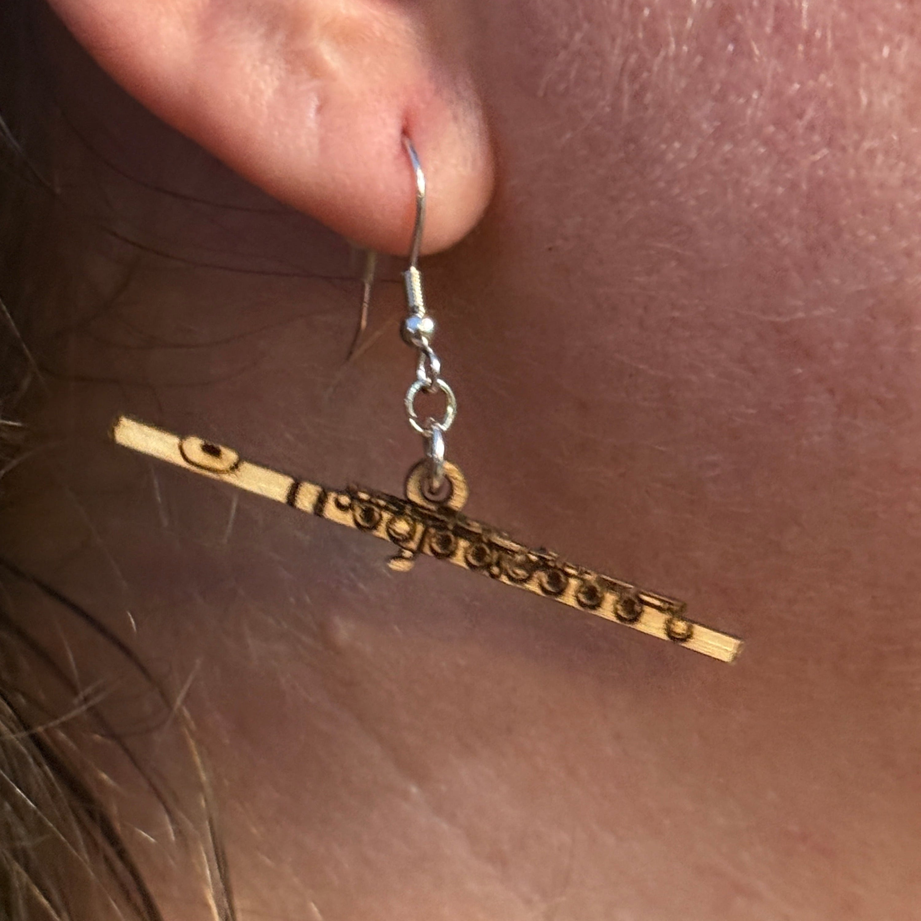 Handmade flute wooden dangle earrings worn on ear, showcasing a detailed laser-cut concert flute design. Lightweight woodwind instrument earrings crafted from wood with hypoallergenic .925 sterling silver hooks, perfect for flutists, band members, orchestra musicians, music teachers, flute students, and music lovers.