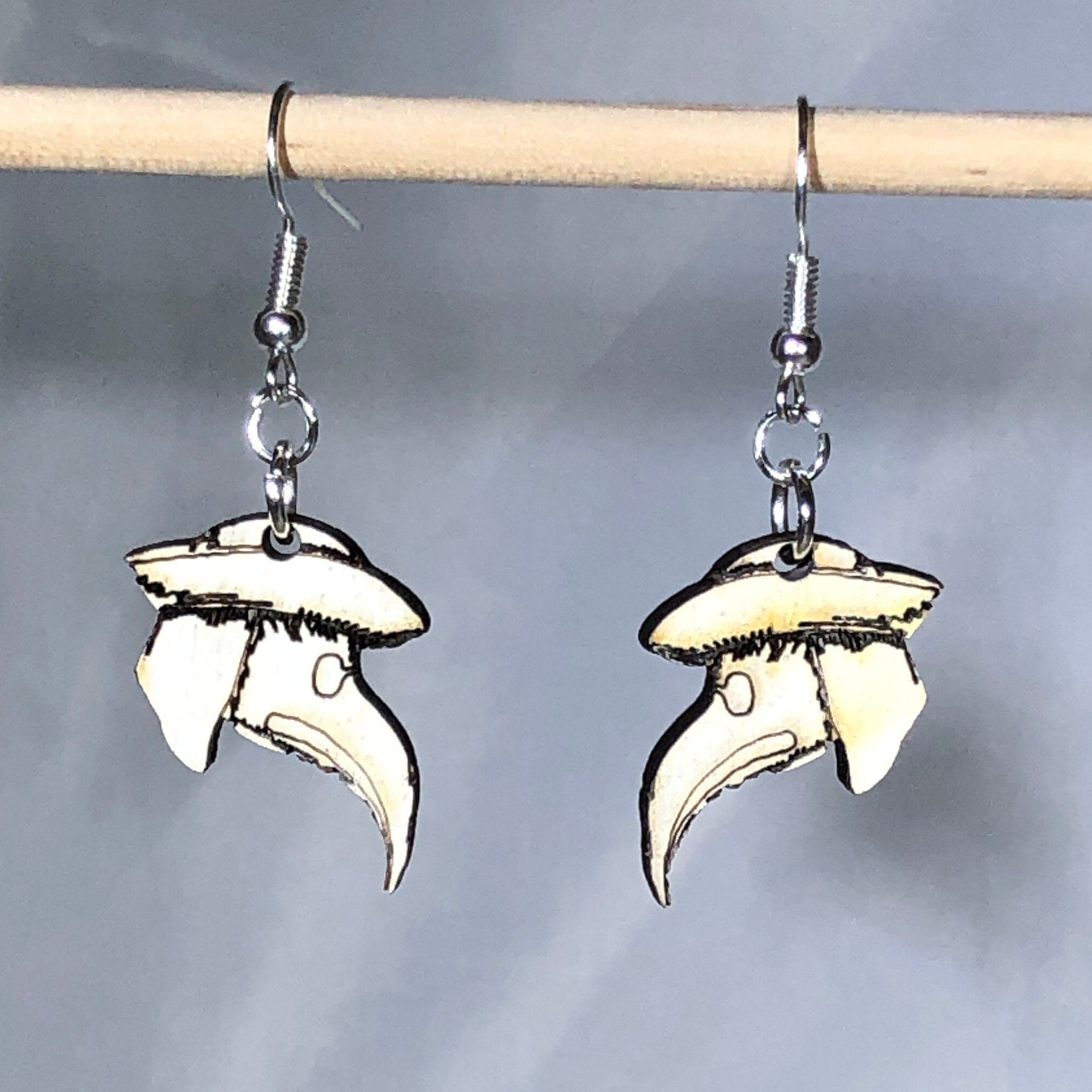 Handmade wooden plague doctor mask dangle earrings with engraved details and a classic beaked mask design on hypoallergenic .925 sterling silver hooks, displayed hanging against a soft blue background. Lightweight gothic jewelry for Halloween lovers, dark academia enthusiasts, horror fans, and historical costume collectors.