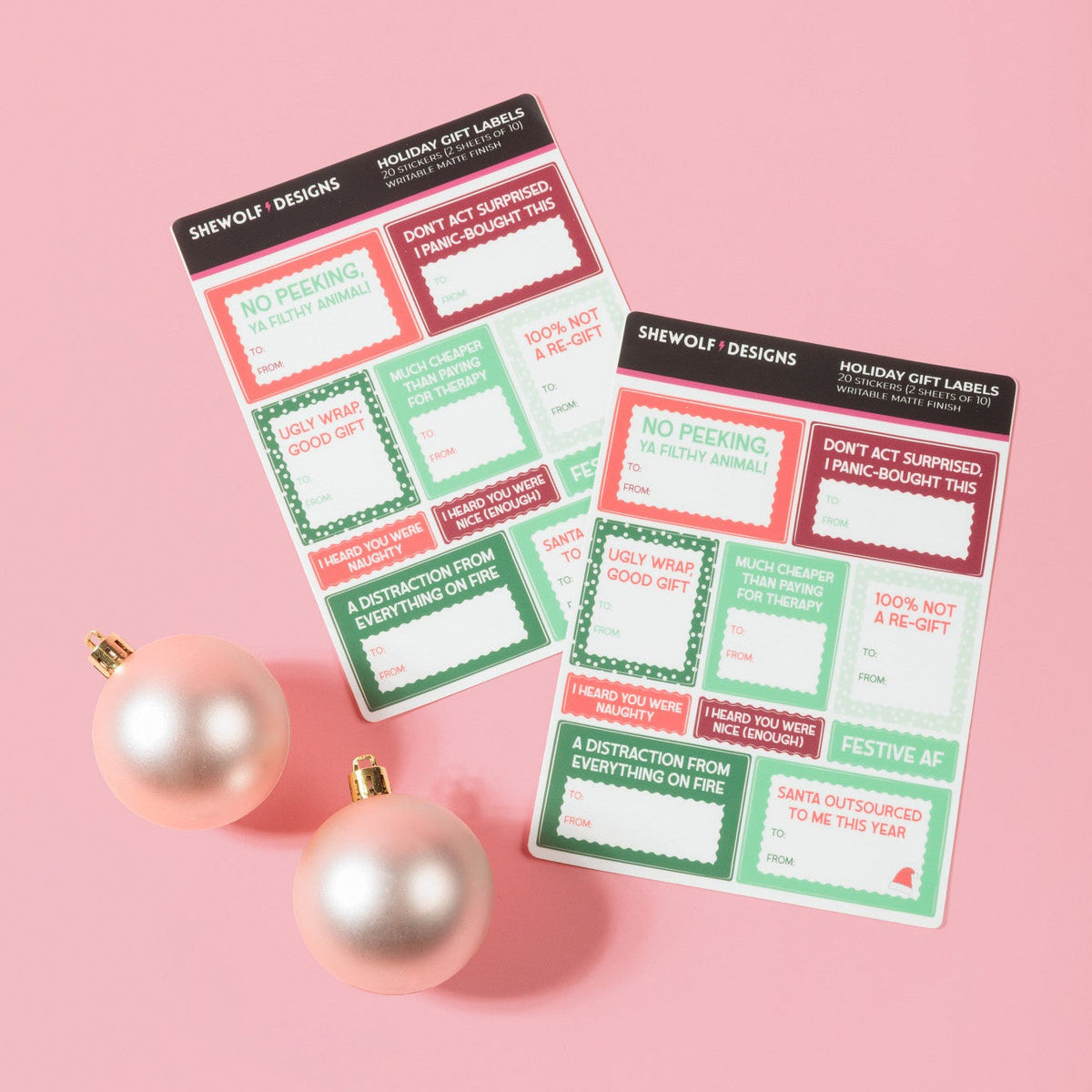 Two holiday-themed gift label sheets from SHEWOLF Designs on a pink background, styled with two pink ornaments. The notepads feature festive red, green, and white holiday labels