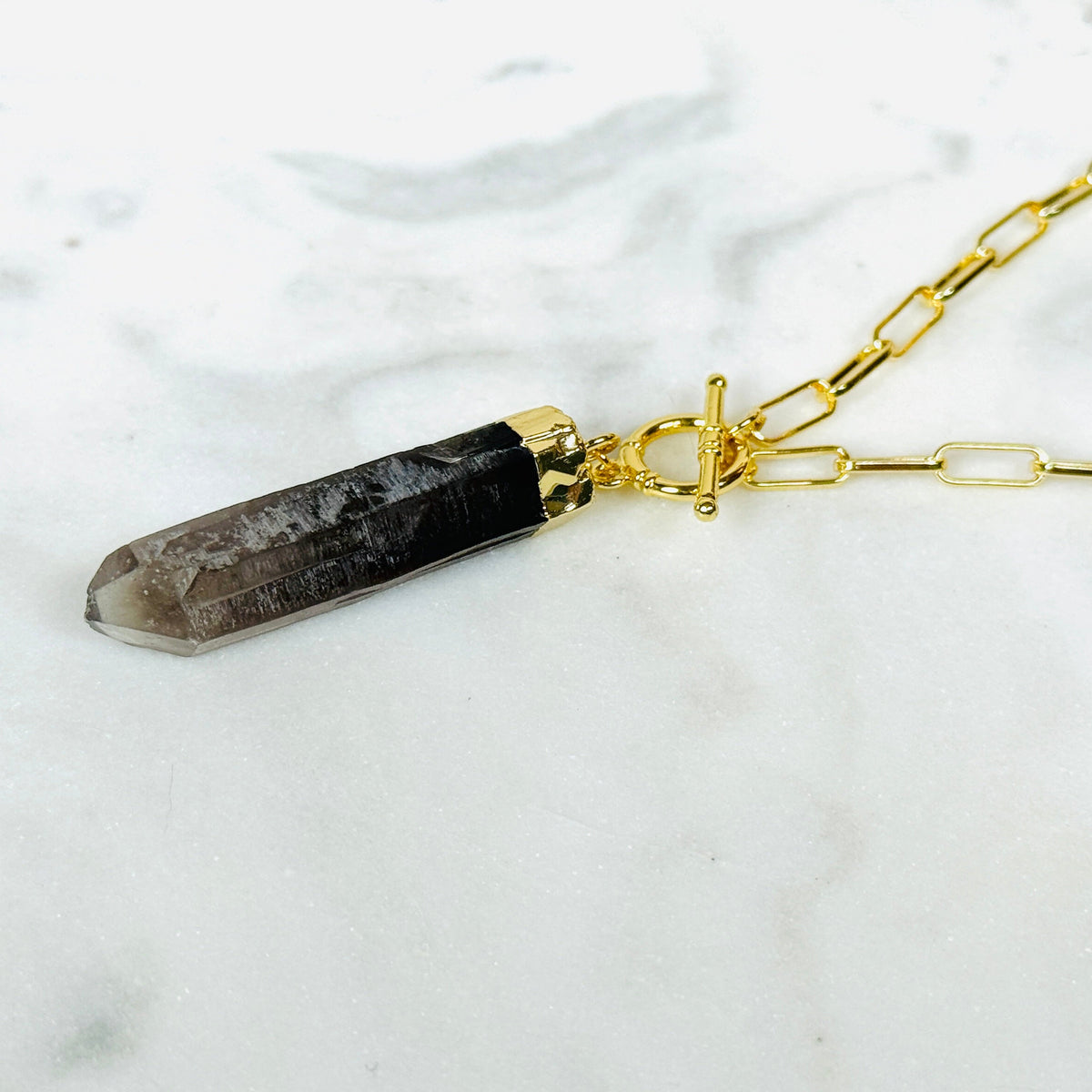 A gold chain necklace featuring a smoky quartz crystal pendant with a gold toggle clasp, displayed on a white marble surface.