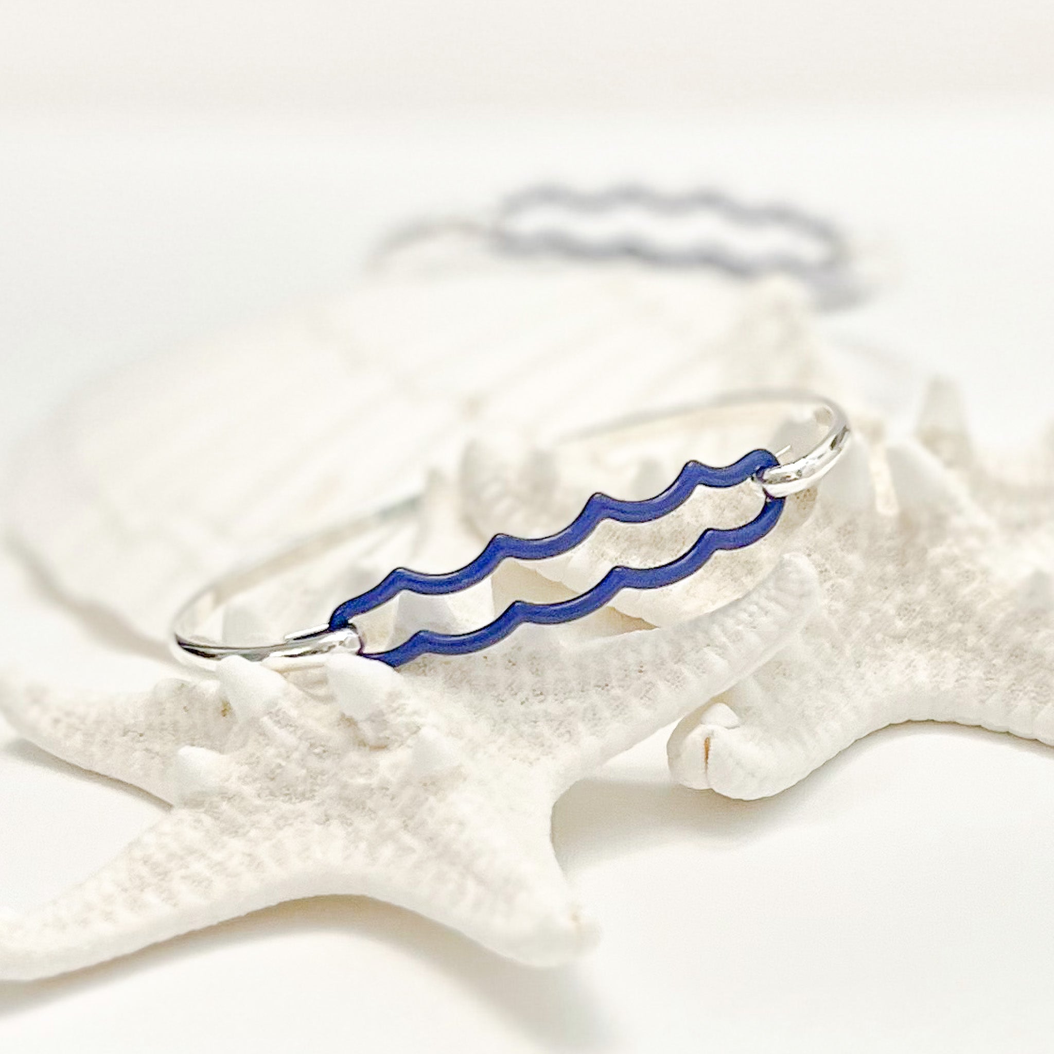 the beach and back double wave tension closure bangle in marine blue enamel with silver bangle set upon white sea stars on white background