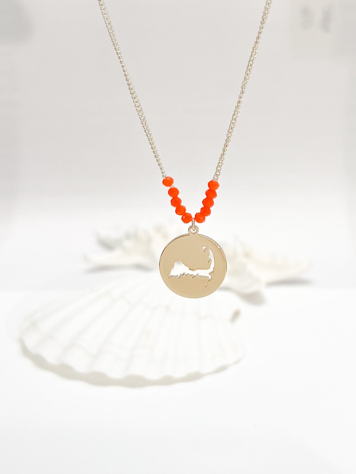 the beach and back cape cod necklace in gold with coral beads at center of chain and coin shape pendant with cutout silhouette of cape cod pictured in front of white seashells on white background