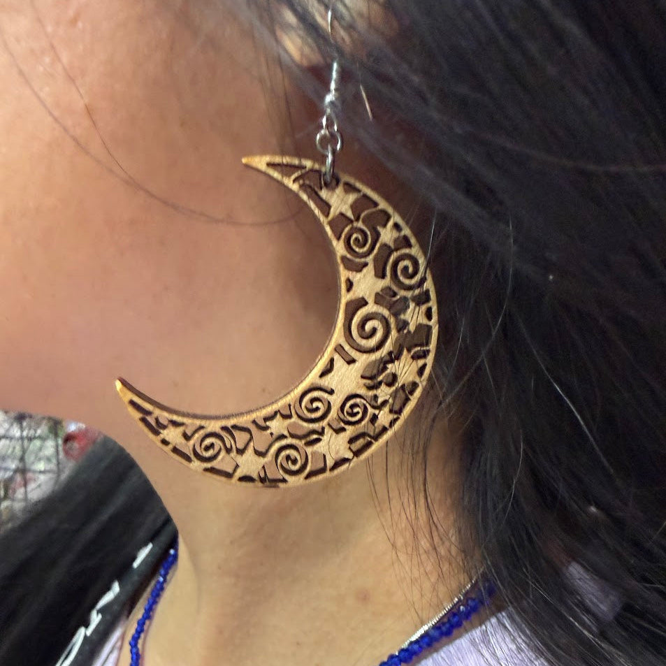 Close-up of wooden crescent moon dangle earring with intricate filigree swirl and star cutout design and hypoallergenic sterling silver hook, celestial boho statement jewelry worn on ear