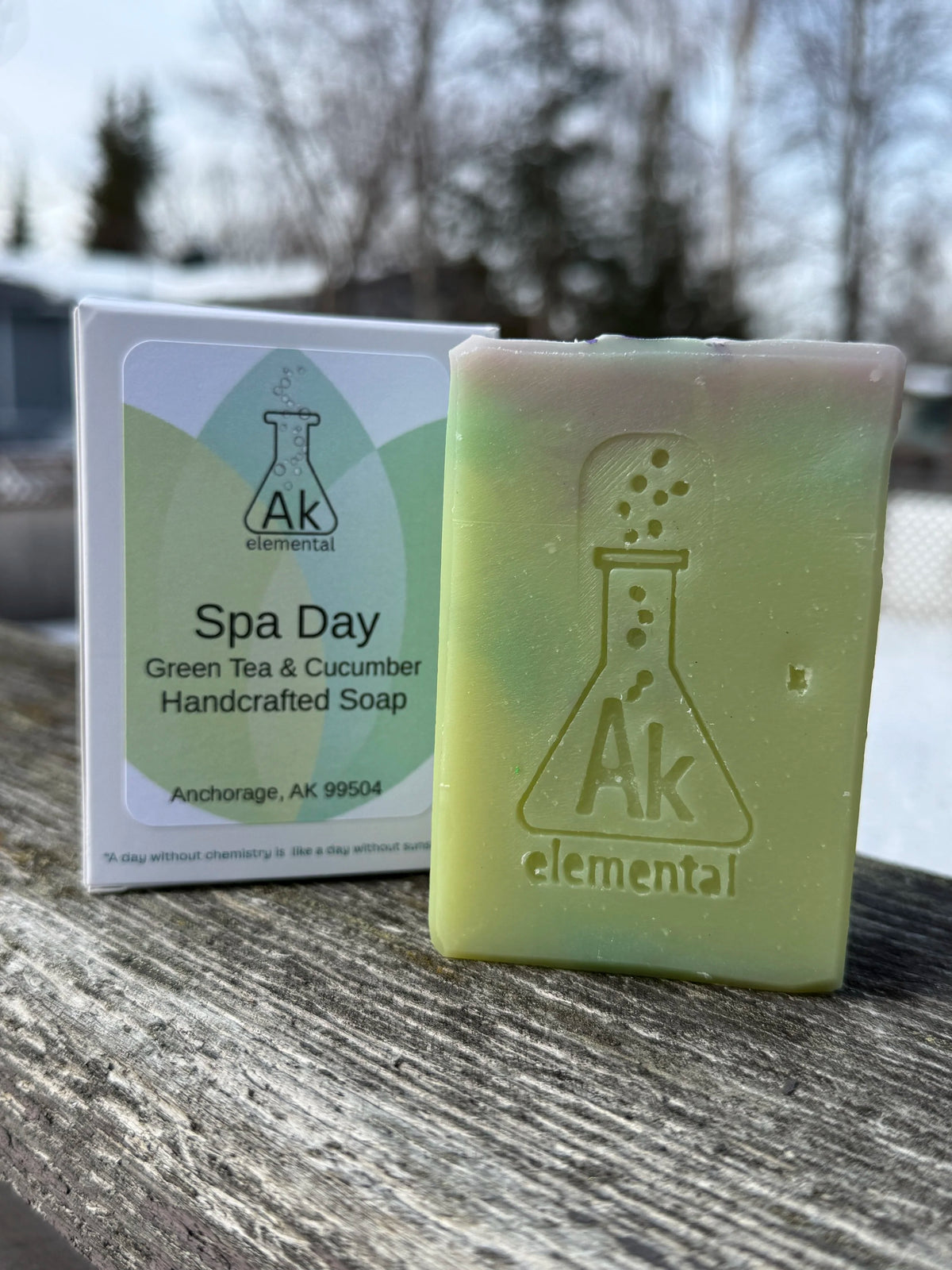 Handcrafted pale green Spa Day soap with AK Elemental logo, packaged beside it on a rustic table with trees in the backdrop.