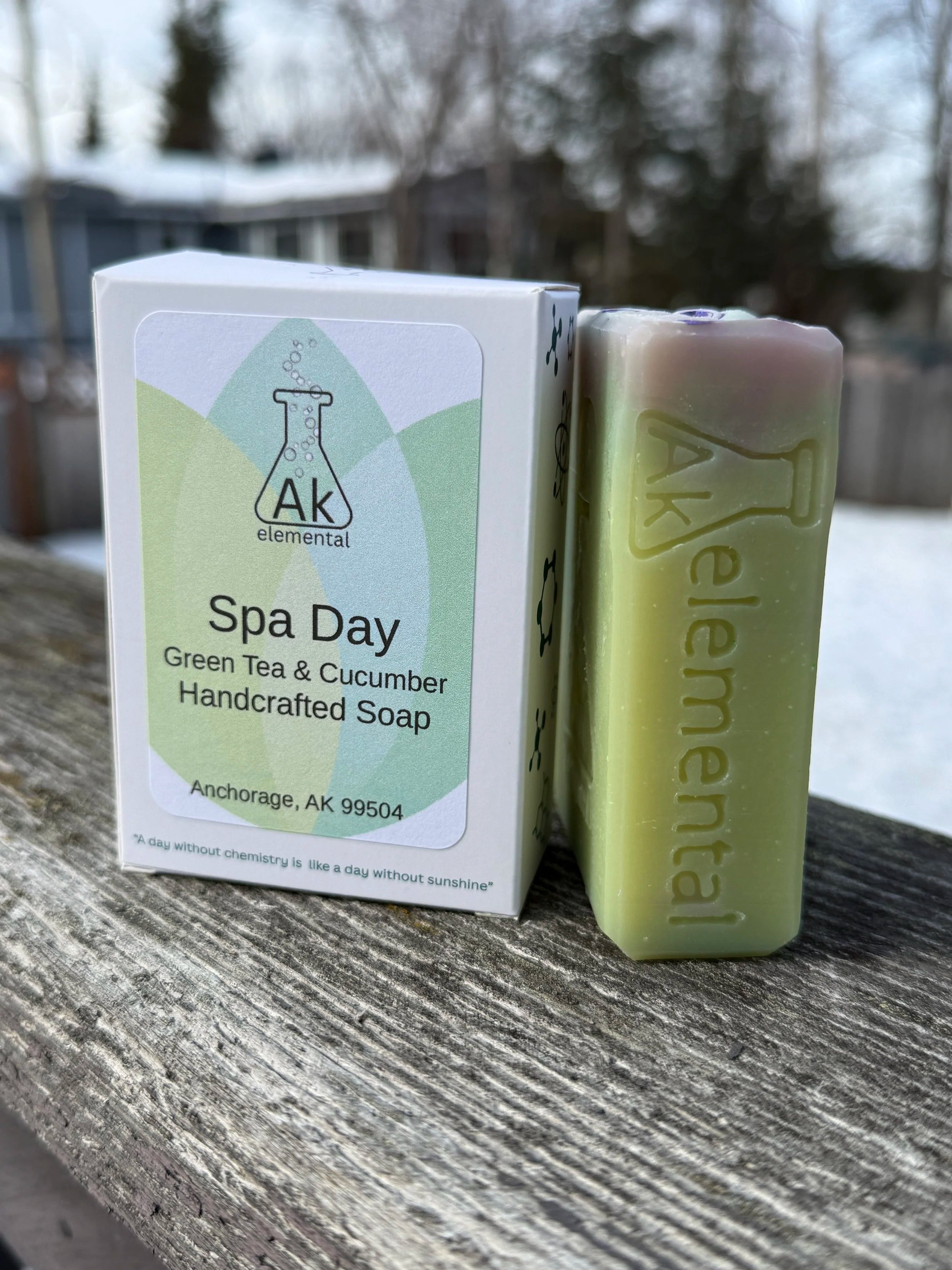 Handcrafted pale green Spa Day soap with AK Elemental logo, packaged beside it on a rustic table with a snowy Alaska backdrop.
