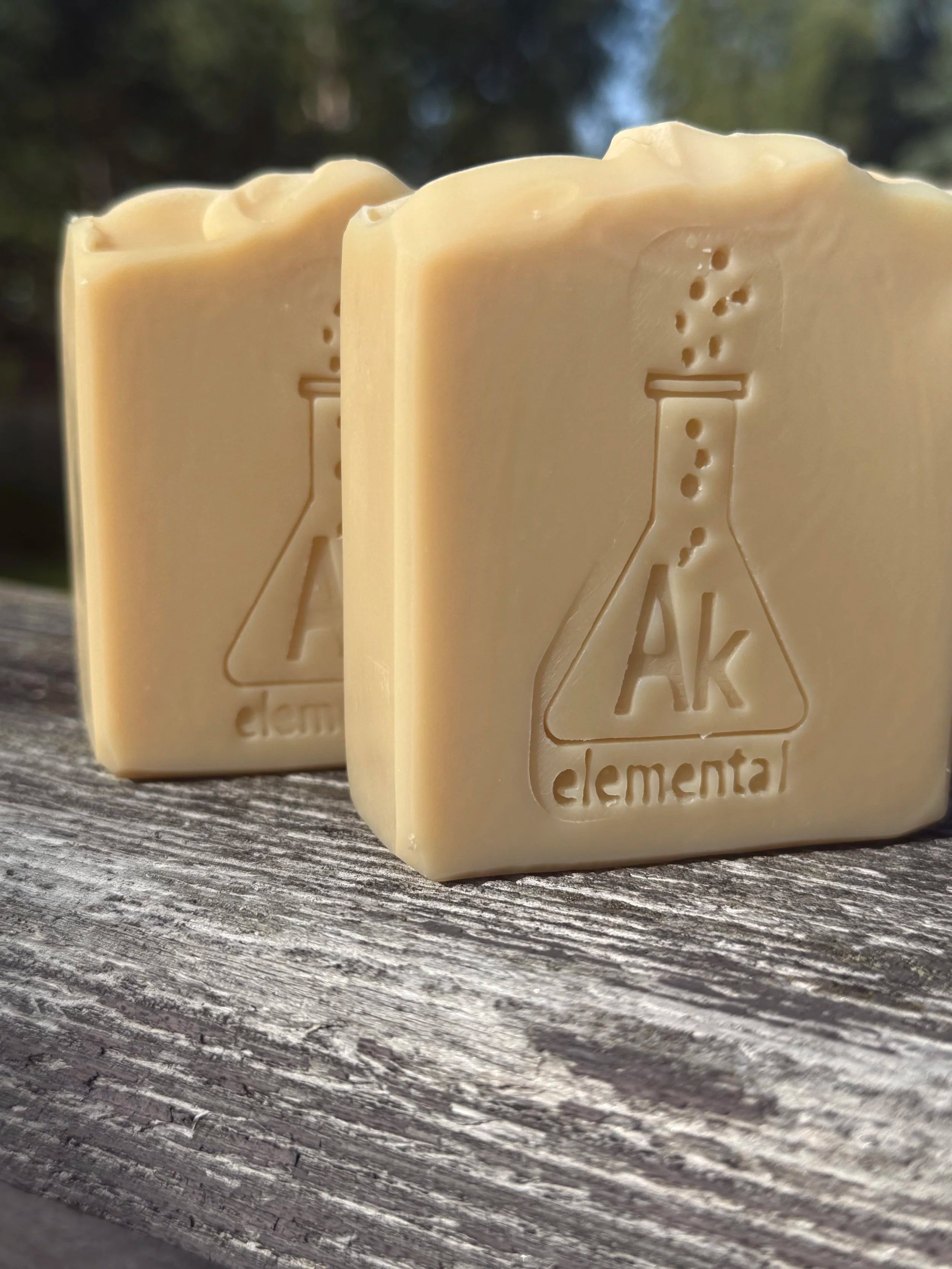 Two creamy, uncolored Sugar Cookie handcrafted soap bars with an embossed AK Elemental flask logo, displayed on rustic wood outdoors.