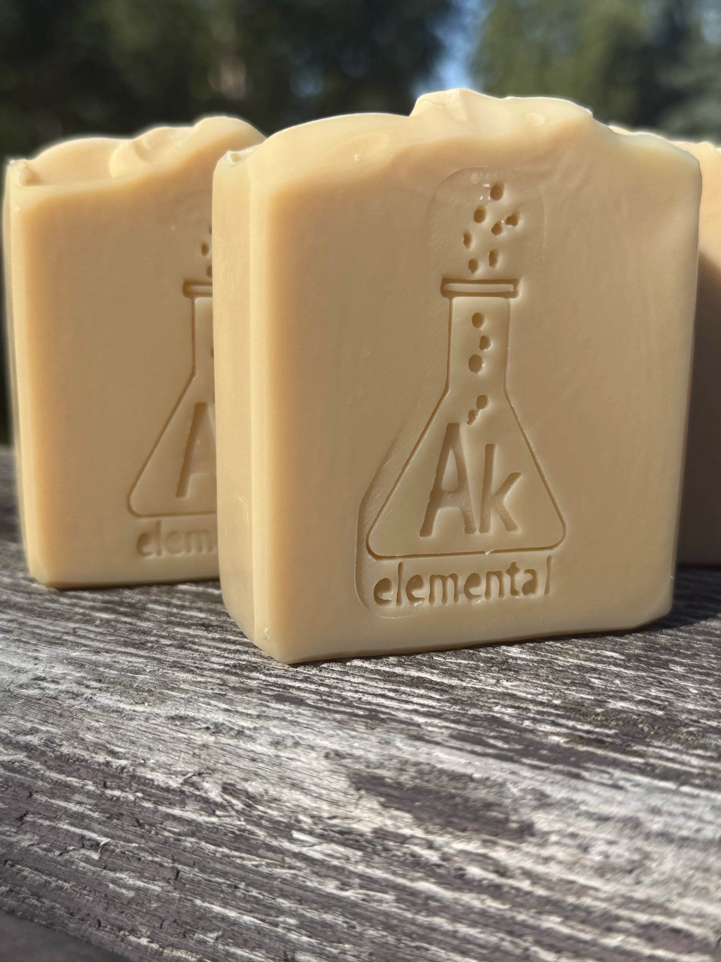 Two creamy, uncolored Sugar Cookie handcrafted soap bars with an embossed AK Elemental flask logo, displayed on rustic wood outdoors.