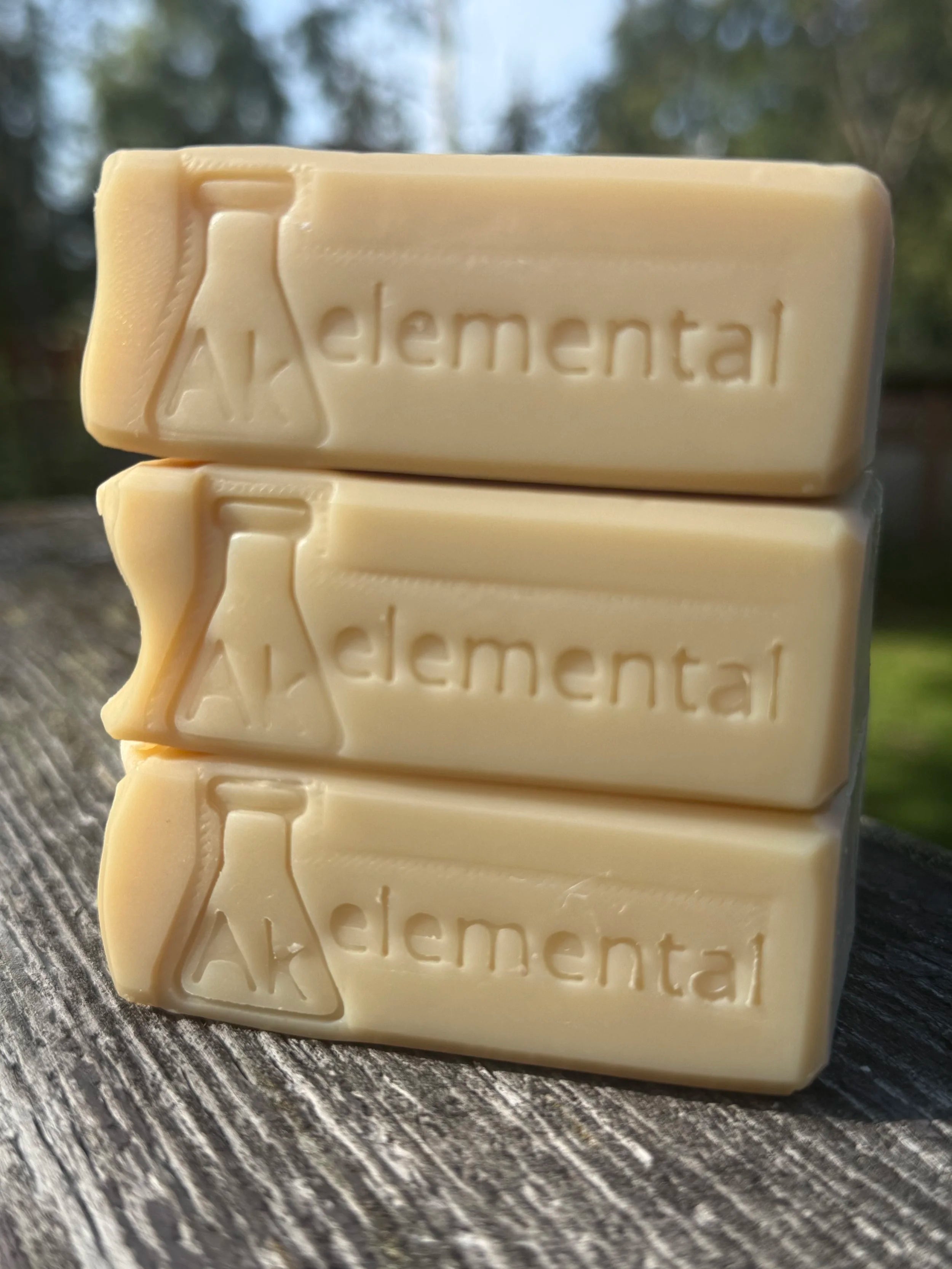 Side view of three uncolored Sugar Cookie handcrafted soap bars with an embossed AK Elemental stamped on the side , displayed on rustic wood outdoors.