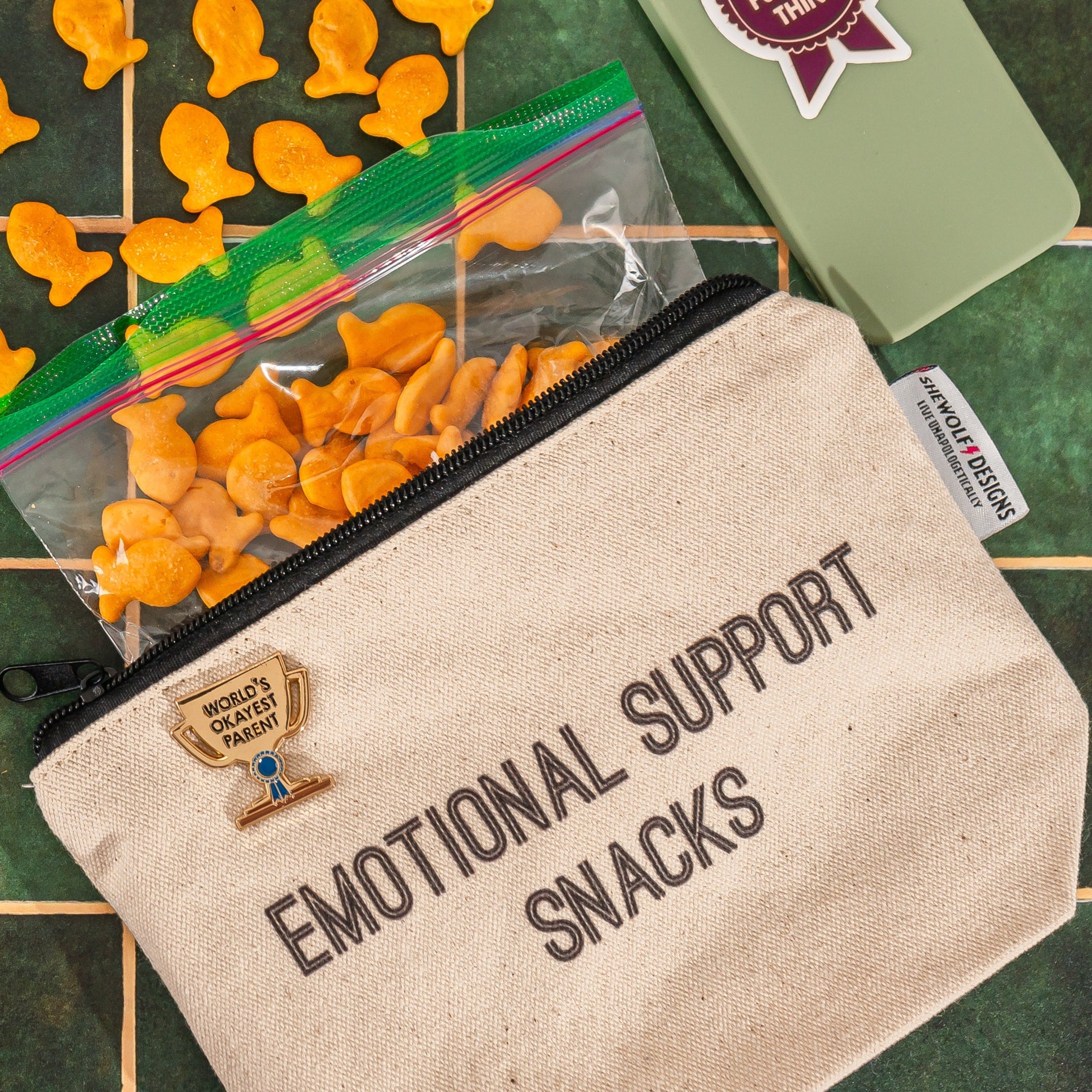 Canvas pouch that says "emotional support snacks" and a gold enamel pin in the shape of a trophy that says "world&