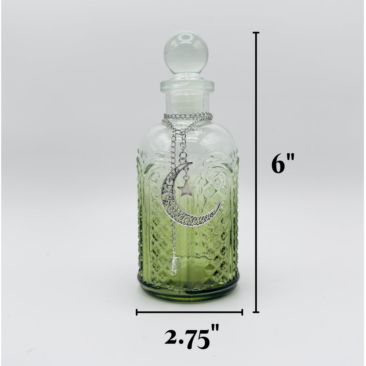 A small clear glass apothecary-style bottle with a stopper and a silver crescent moon pendant, shown on a light gray background with dimension labels indicating 6 inches tall and 2.75 inches wide.