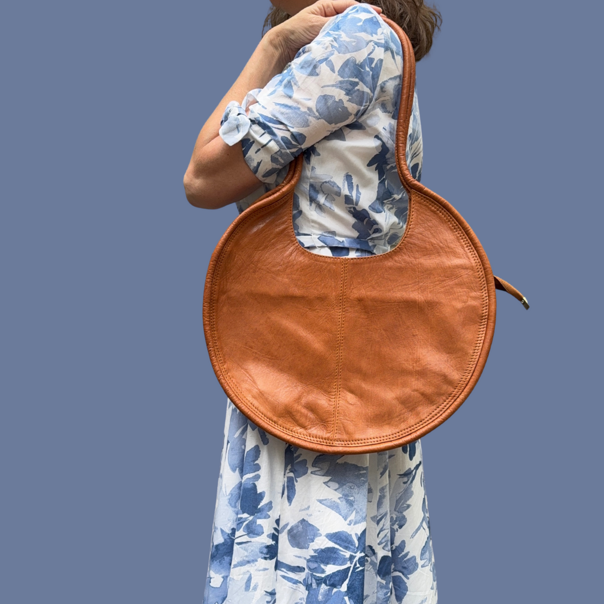 A person wearing a floral dress is seen from the back, carrying an natural leather circular purse over their shoulder.
