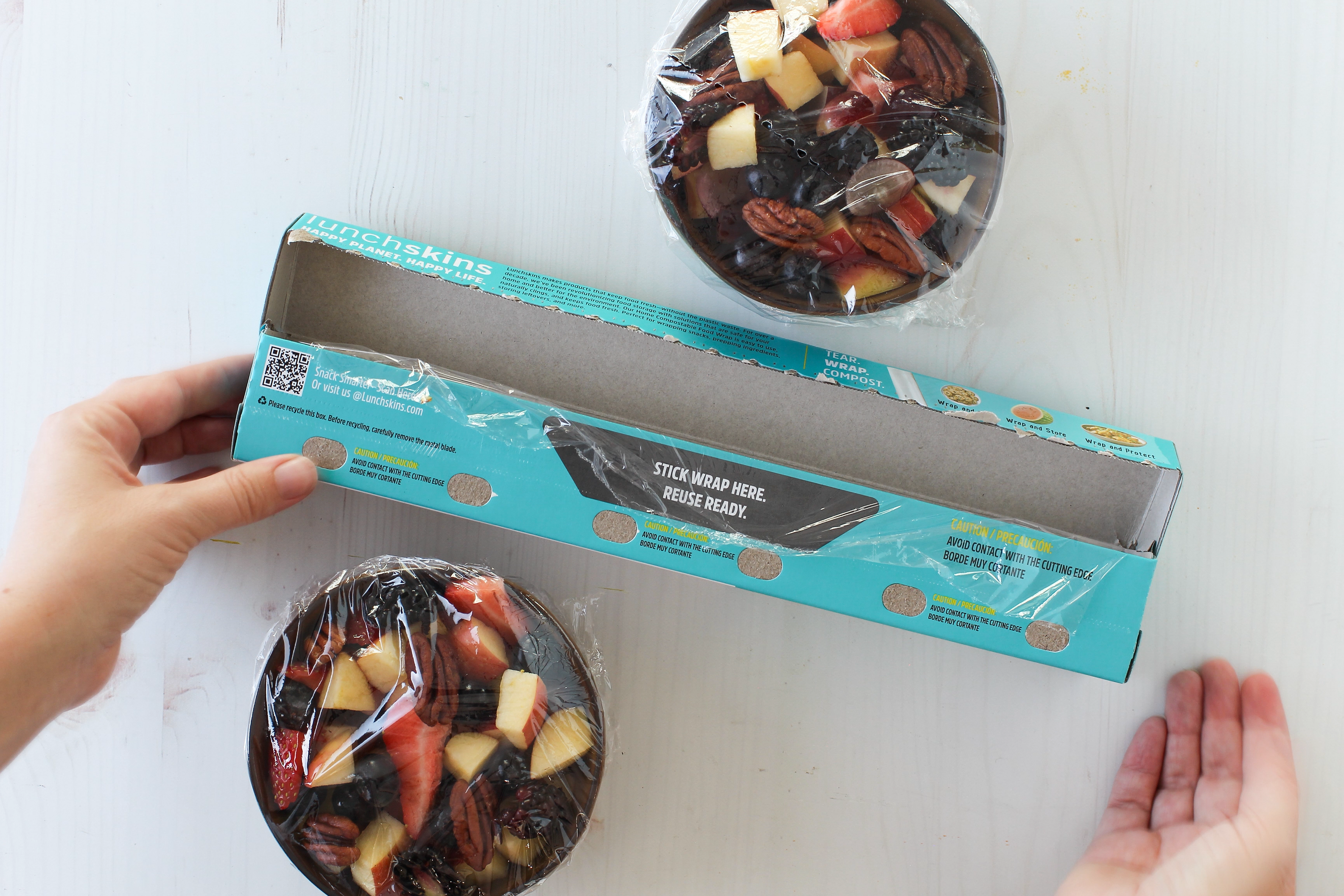 Hands opening a box containing a fruit with a visible brand logo. Showing Reuse Ready Tab Lunchskins Food Wrap