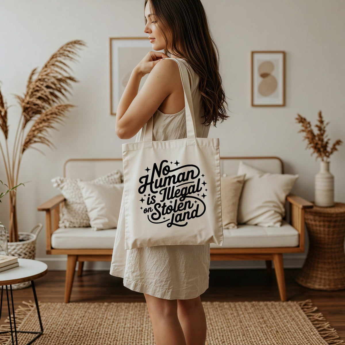 woman holding canvas tote bag that reads no human is illegal on stolen land in a retro script font