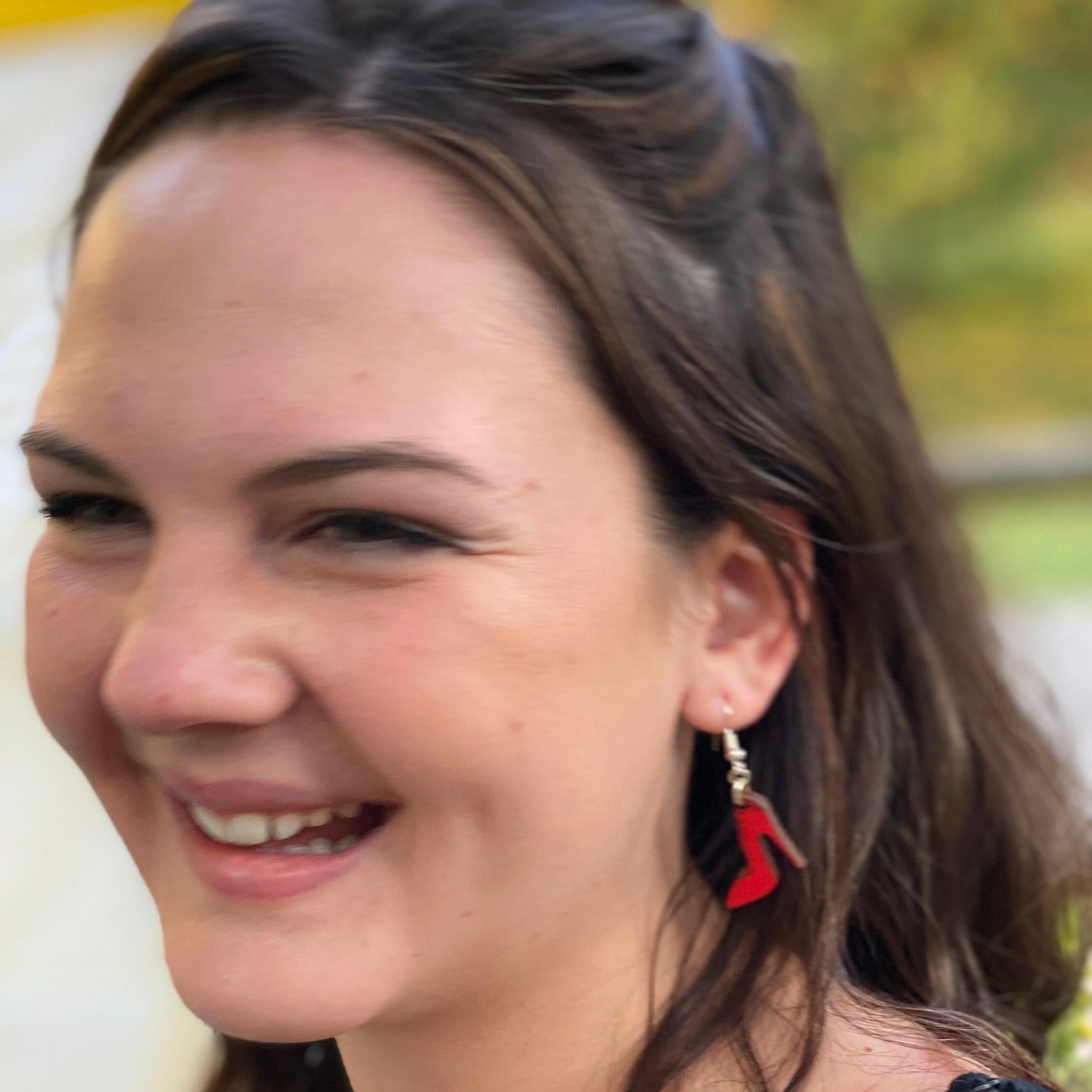 Person wearing hand-painted red high heel wooden dangle earring on a hypoallergenic .925 sterling silver hook, showing the lightweight size and fit when worn. Fashion shoe jewelry for boutique style, shoe lovers, and bold statement accessories.