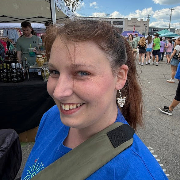 Smiling woman wearing hand-painted wooden pitbull face dangle earrings at an outdoor festival booth, lightweight dog lover pet jewelry by Cate’s Concepts