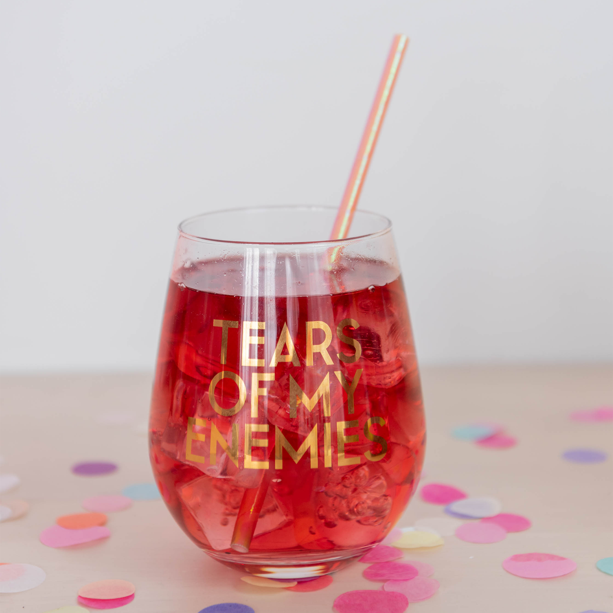 "Tears of My Enemies" stemless glass with a red drink and a striped straw amid pastel confetti.
