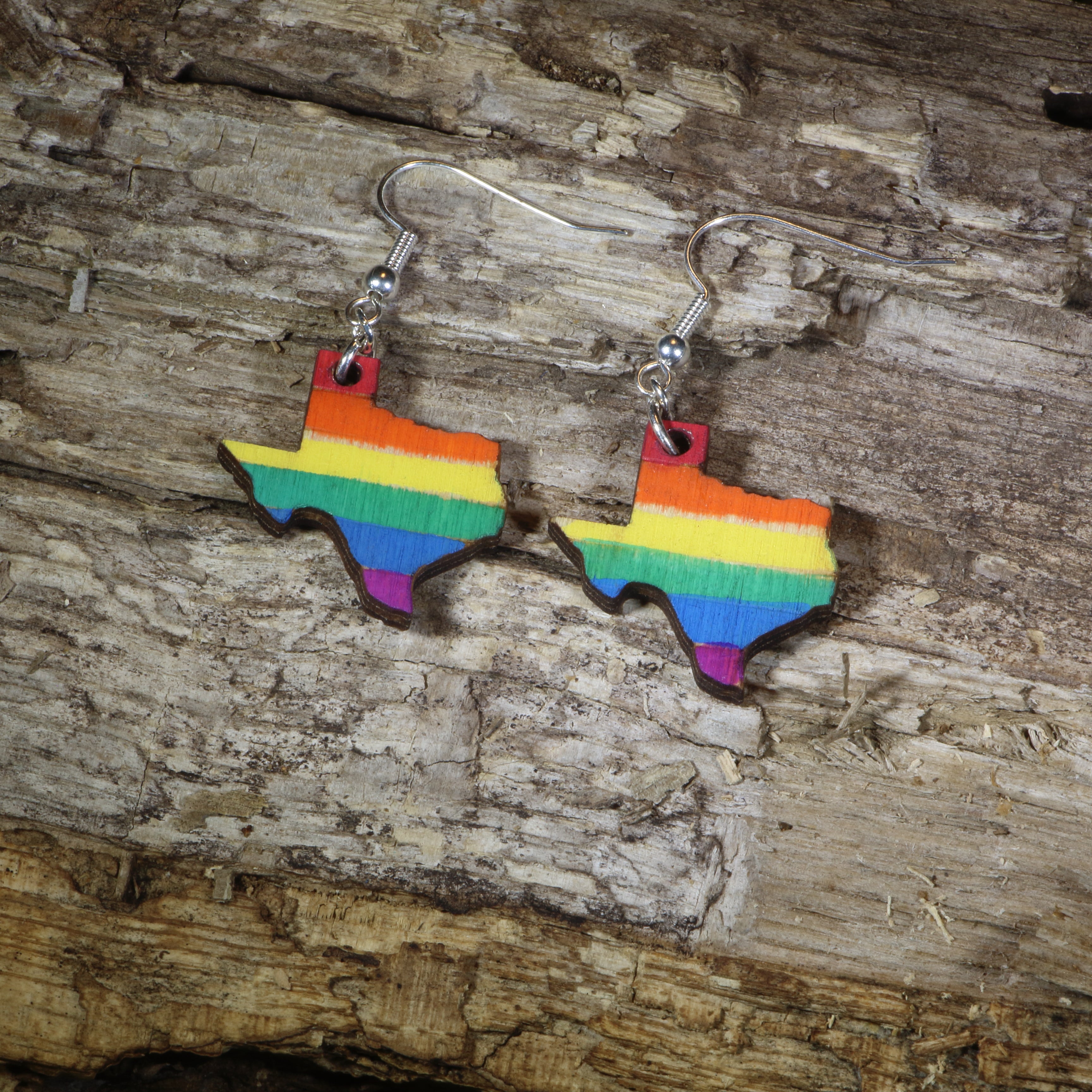 Hand-painted wooden Texas LGBTQ Pride dangle earrings featuring the Texas state silhouette with vibrant rainbow pride stripes, hypoallergenic sterling silver hooks, and lightweight LGBTQ+ state pride jewelry by Cate&