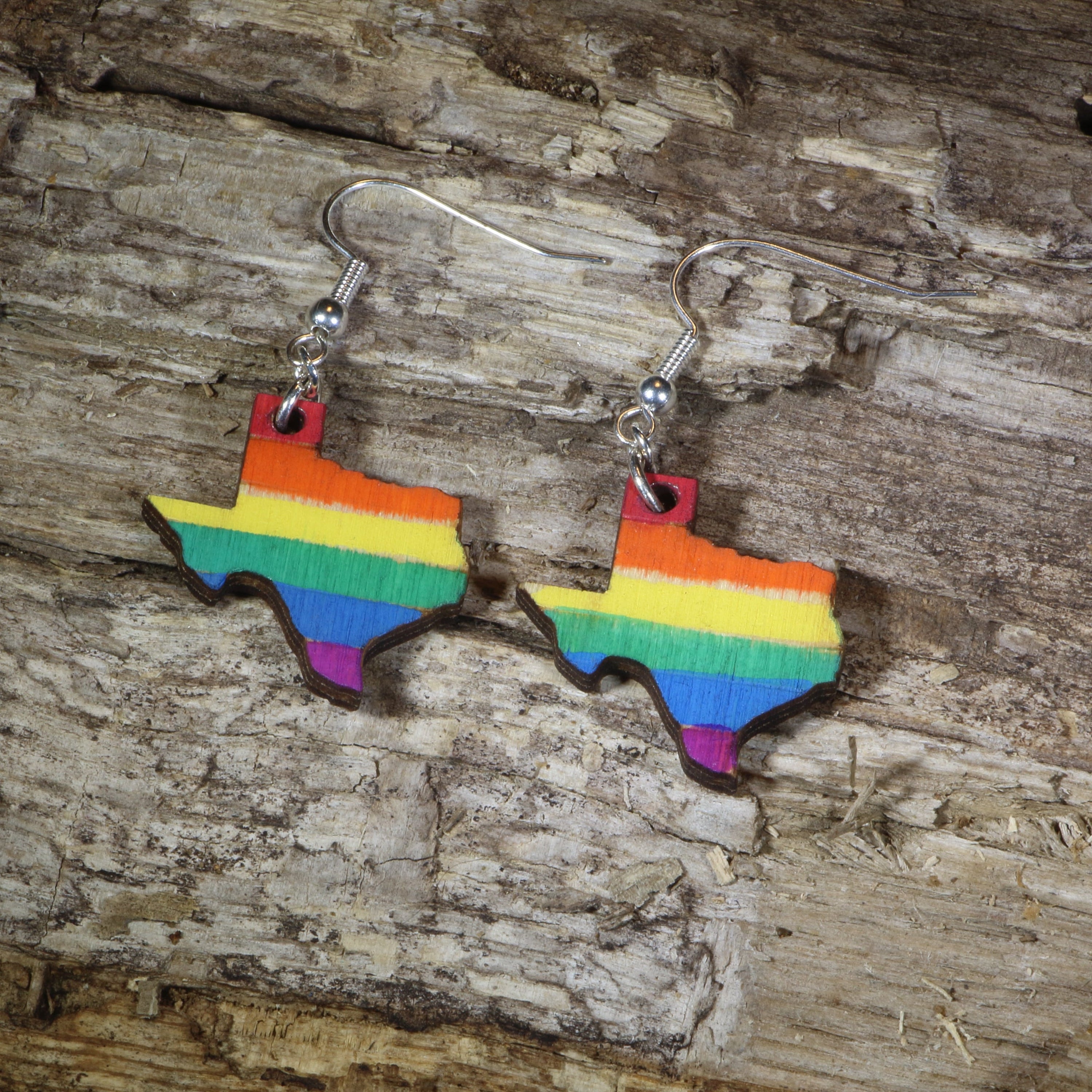 Hand-painted wooden Texas LGBTQ Pride dangle earrings featuring the Texas state silhouette with vibrant rainbow pride stripes, hypoallergenic sterling silver hooks, and lightweight LGBTQ+ state pride jewelry by Cate&