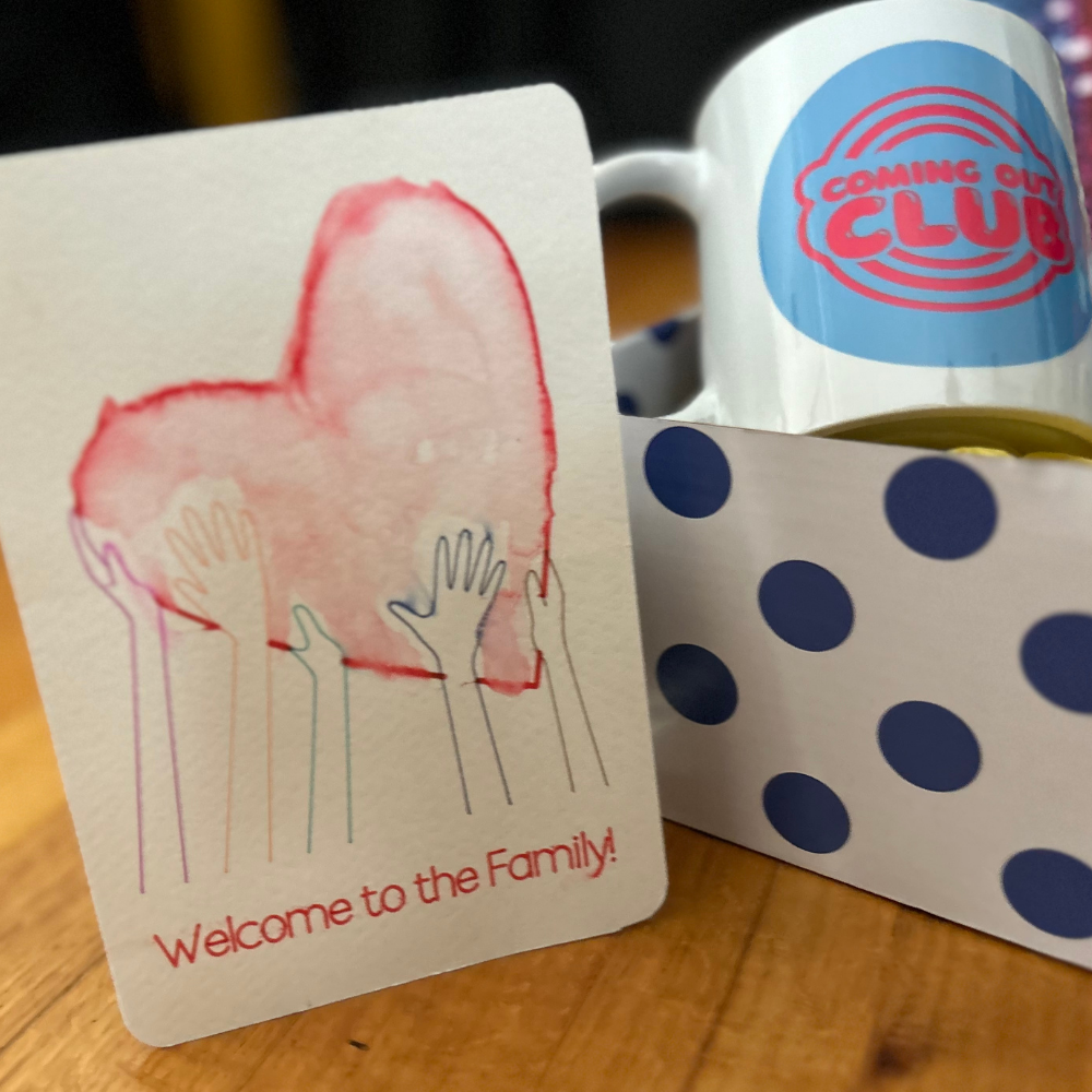 The contents of the box are inside a blue and white spotted box. On the lef is the Welcome to The Family postcatd (with a watercolor red heart) Inside the box is a coming out club mug with blue/white background and red letters