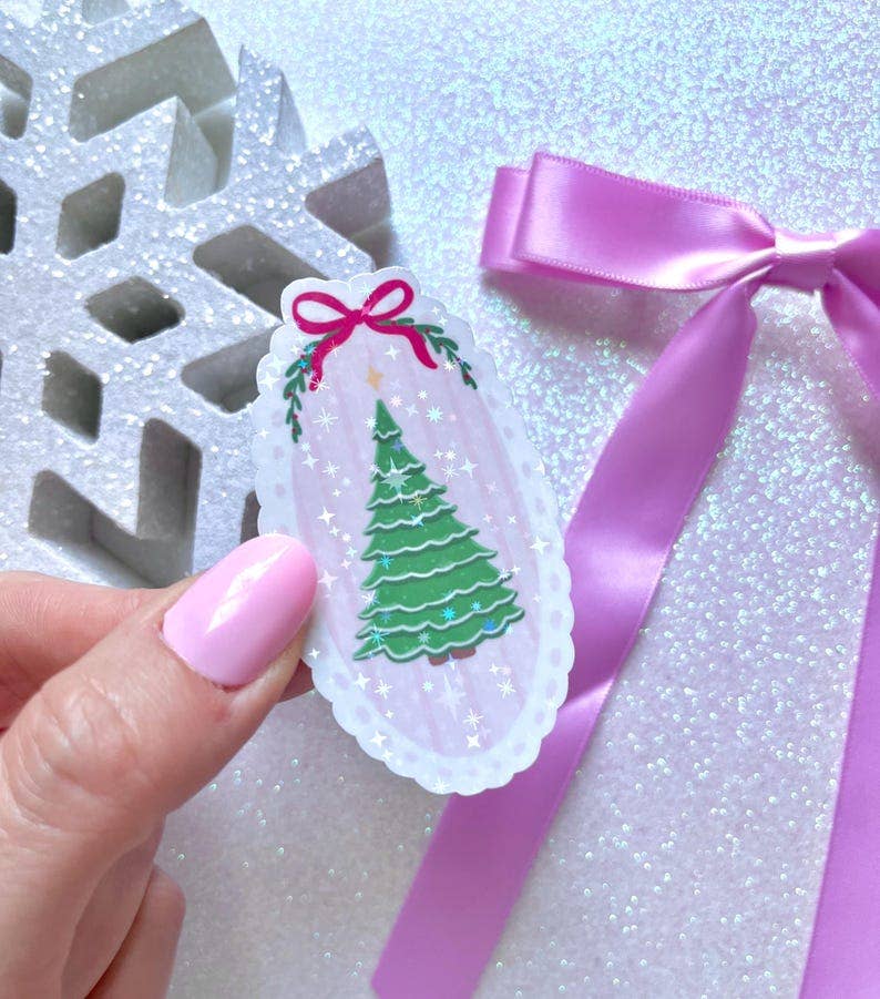 A hand with pink nail polish holds a small oval ornament or sticker featuring a green Christmas tree, white scalloped border, and pink bow, against a glittery light background with a purple ribbon and a white snowflake decoration.