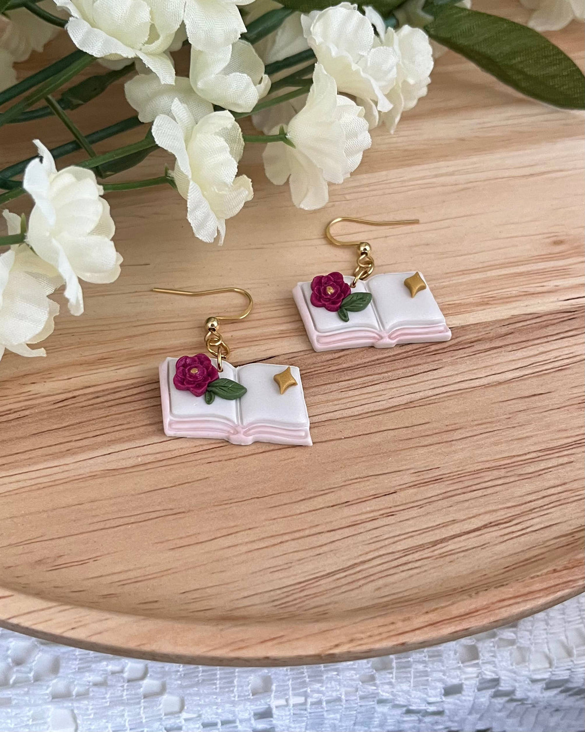 Book-shaped earrings with floral details on a wooden surface with white flowers.