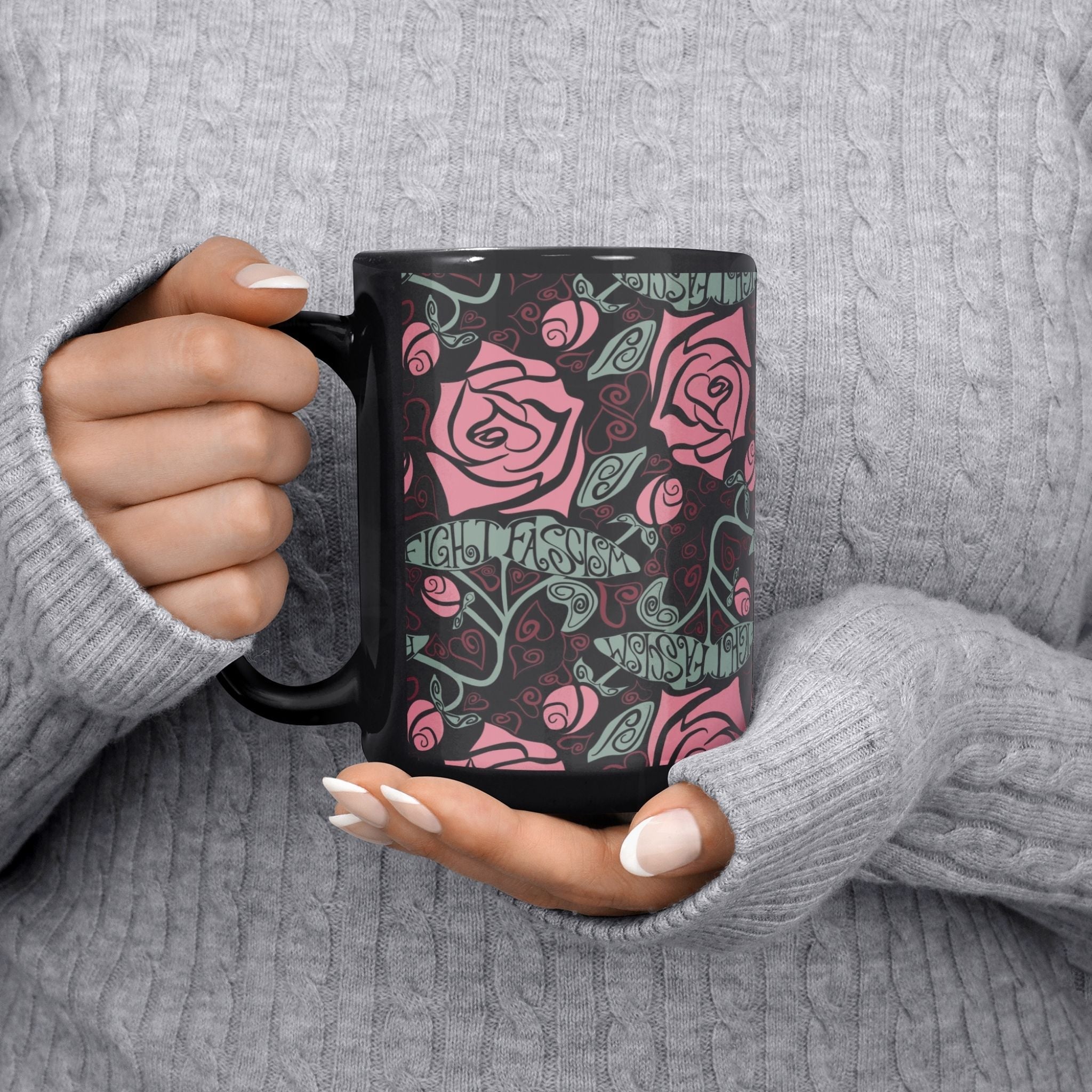 Both hands cradling the Fight Fascism anti-fascist mug in a cozy sweater, warm pink rose and vine stem wrap print on black ceramic